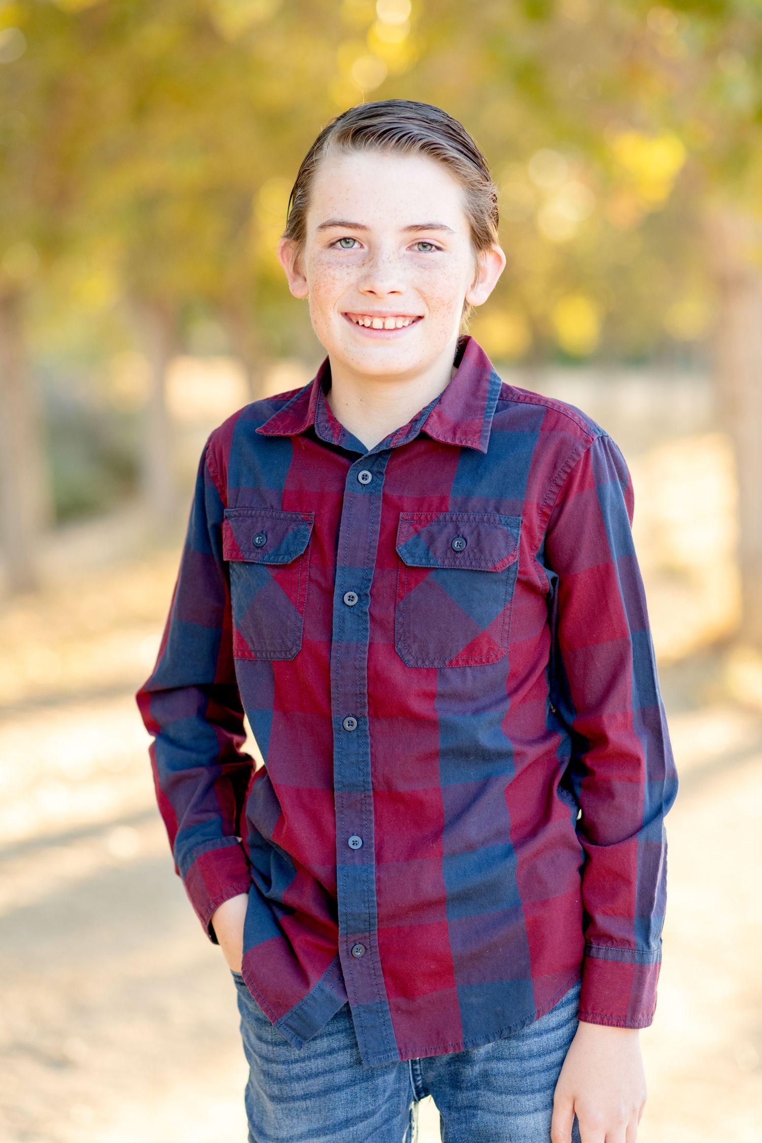 Child wearing red and blue plaid button-up shirt standing outdoors in autumn setting.