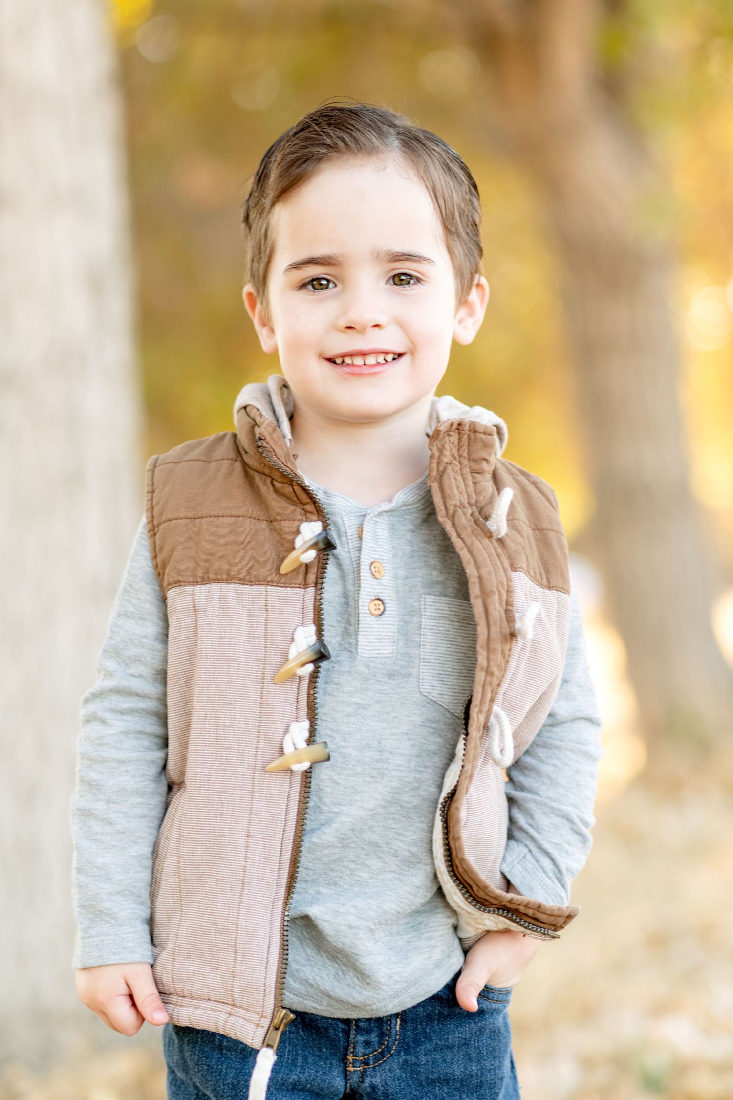 Child wearing brown vest smiles at camera during autumn outdoor portrait session.