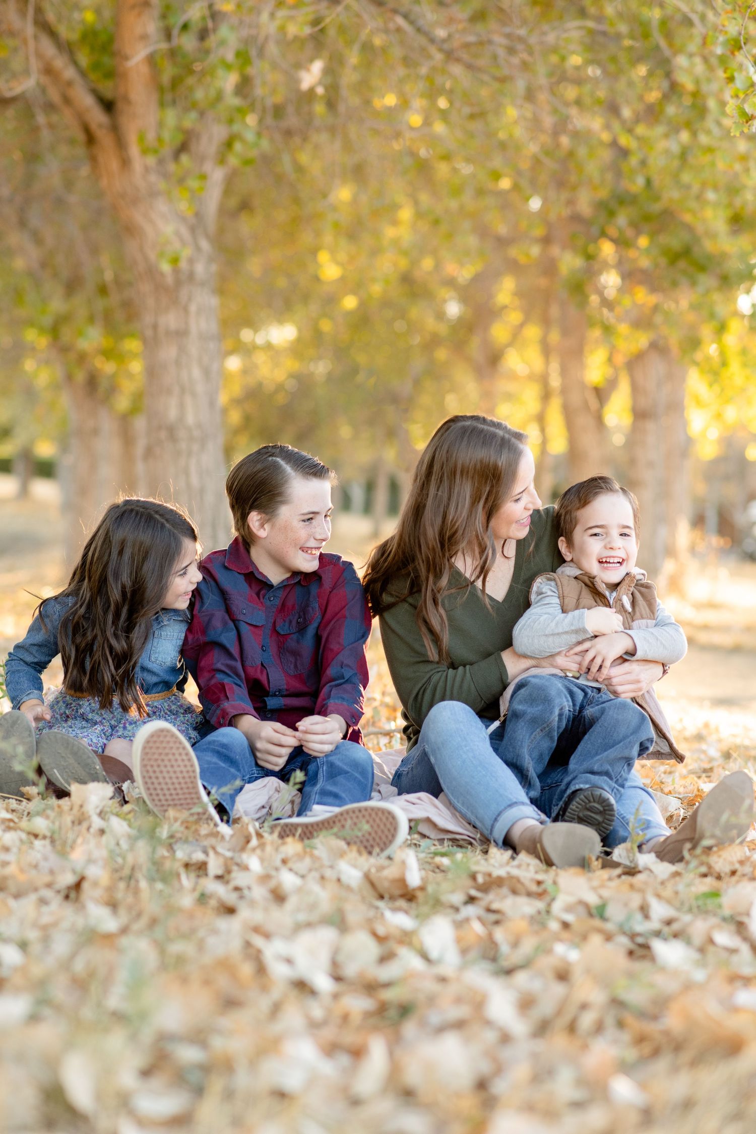 Family sits together on ground covered with fallen autumn leaves under golden trees.