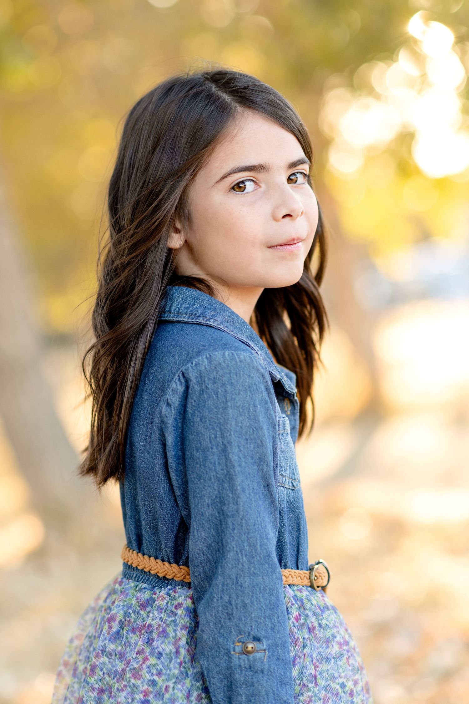 Child in denim jacket and floral dress poses against golden autumn background.