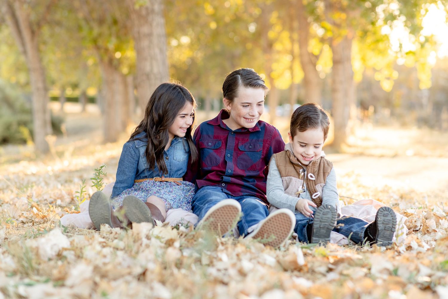 Three children sit together laughing on ground covered with autumn leaves.