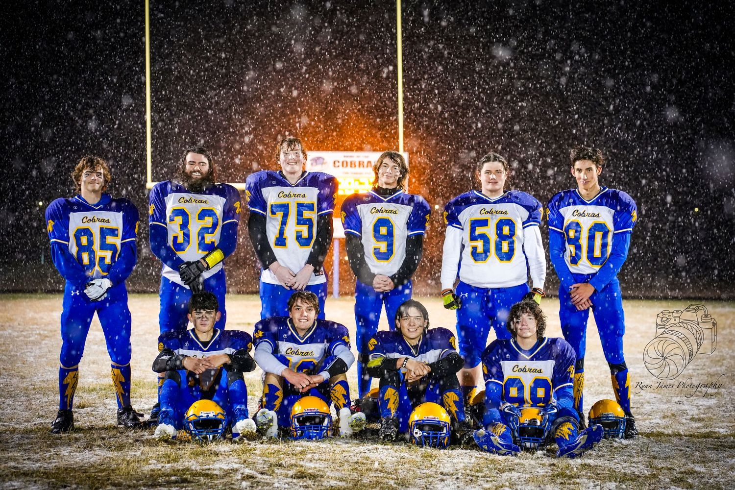 Football team poses together in blue uniforms during a snowy night game under bright stadium lights.