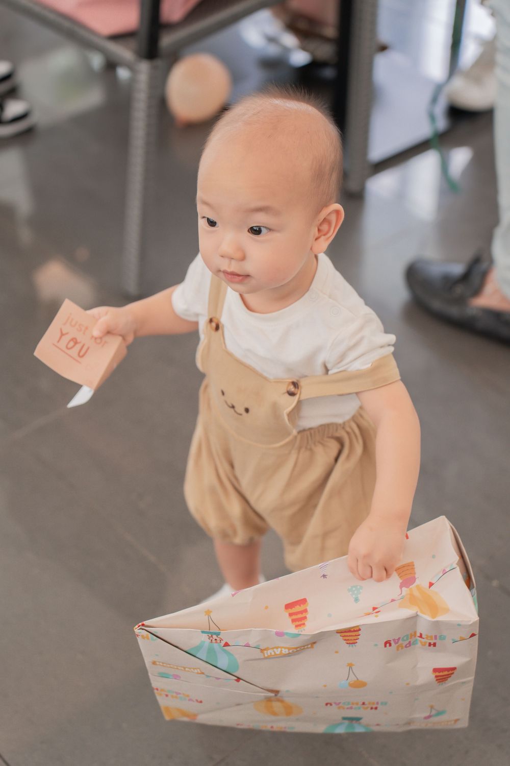 Toddler in tan romper plays with decorated paper box on floor.
