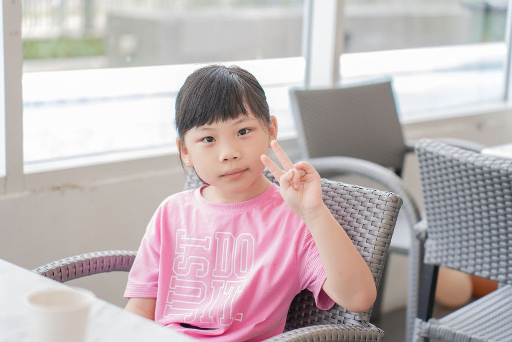 Child in pink t-shirt makes peace sign while seated at cafe table.