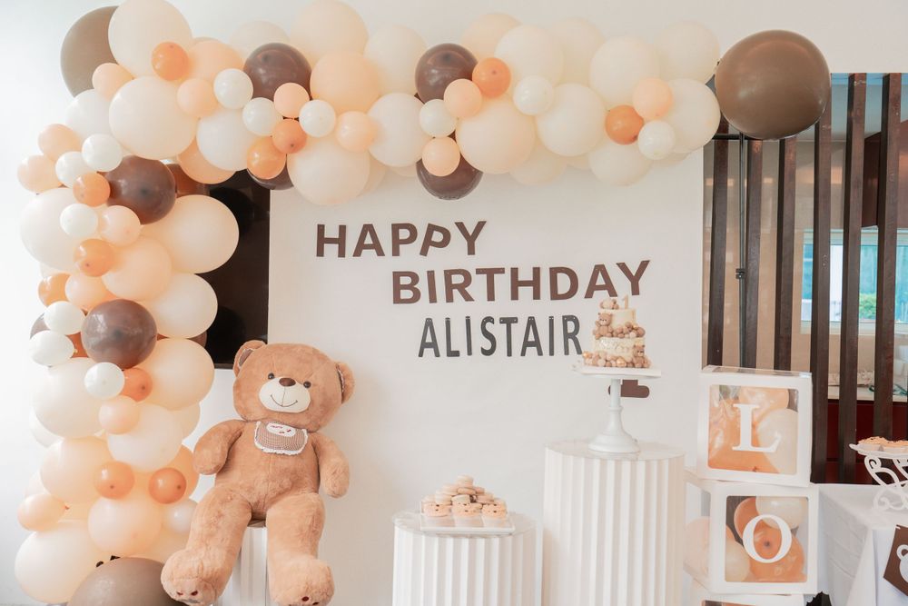 Birthday party decorations with balloon arch and teddy bear theme in peach and brown colors.