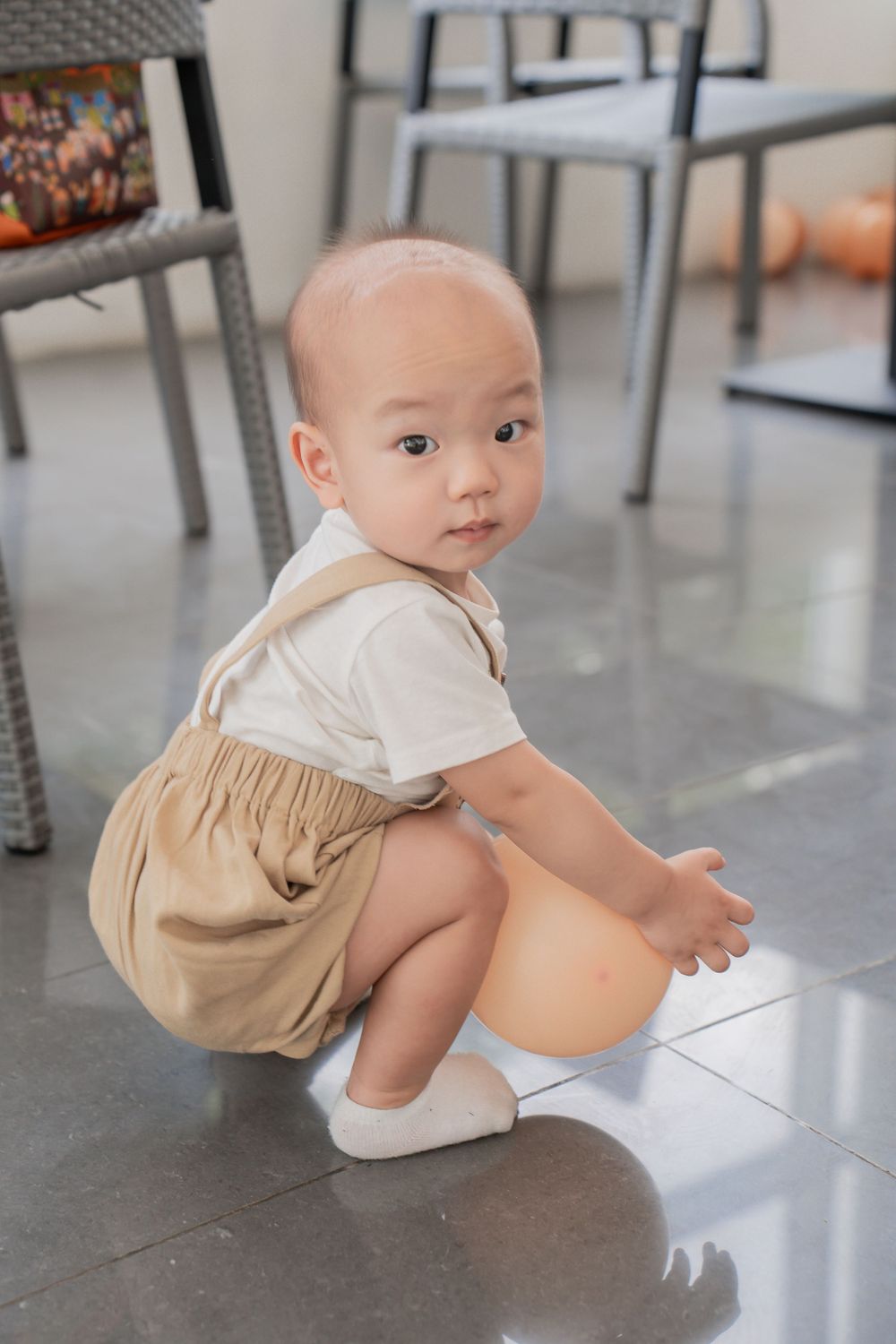Young toddler in beige overalls and white shirt crouches on reflective floor.