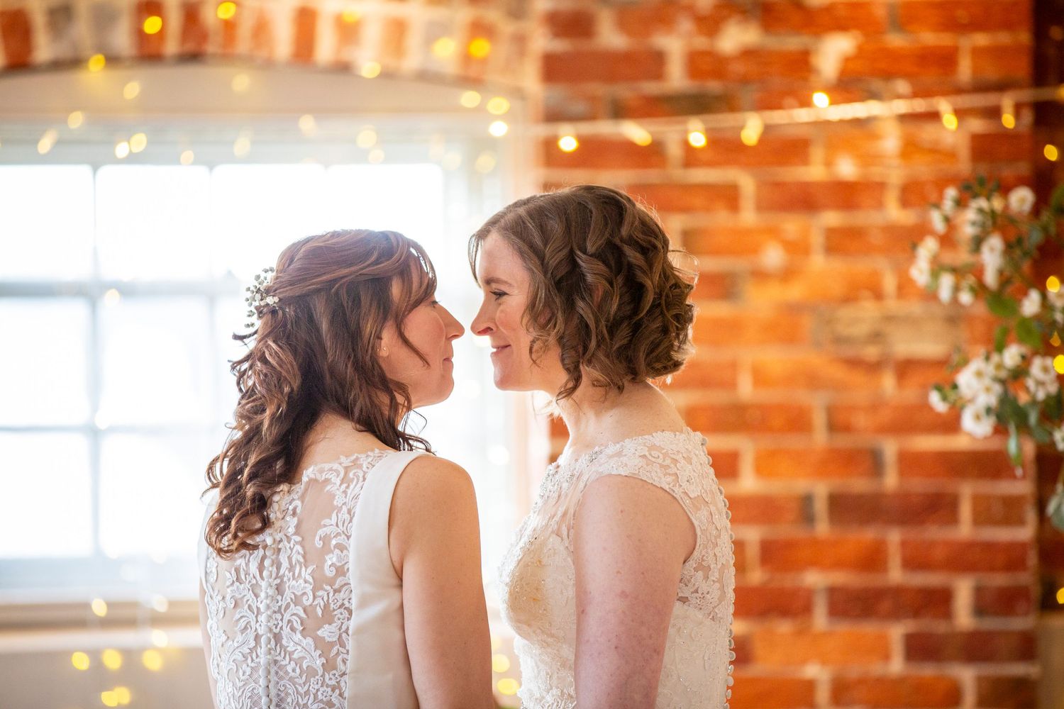 Two brides share an intimate moment in front of a rustic brick wall with string lights.