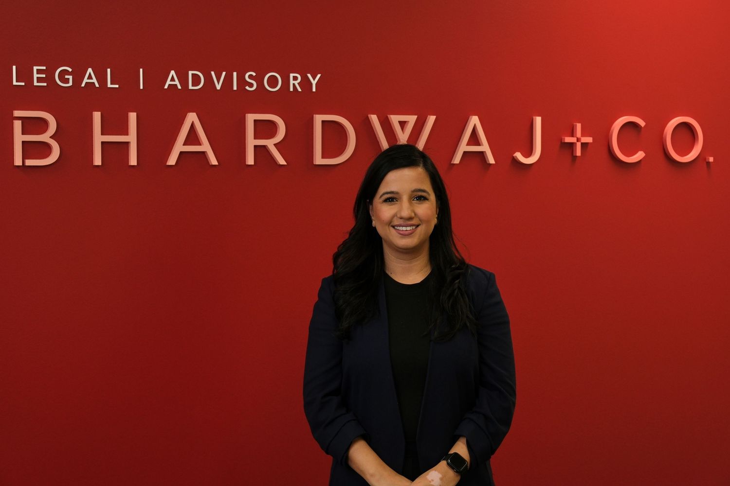 Professional woman stands in front of red Bhardwaj Co wall signage wearing business attire.