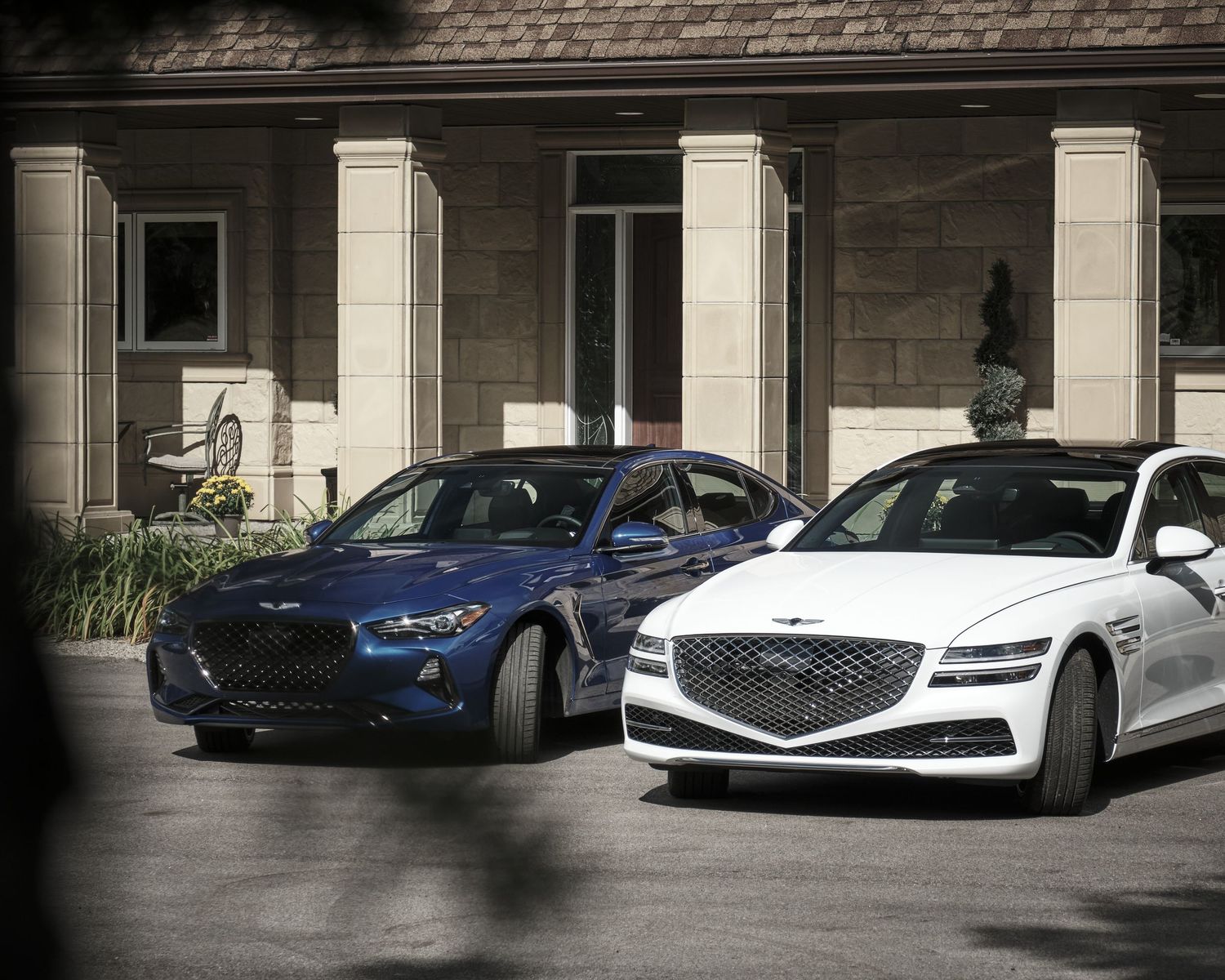 Two luxury Genesis sedans, one blue and one white, parked in front of a house with white columns.