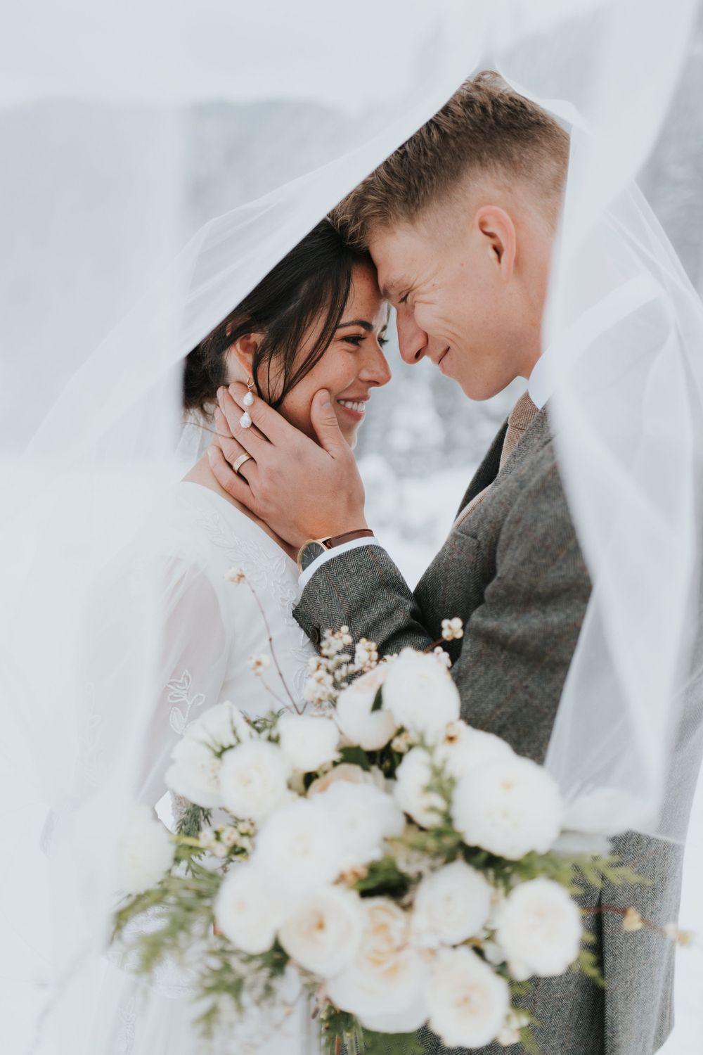 A bride and groom smile underneath a veil as they take their wedding portraits in the snowy mountains of Provo, Utah.