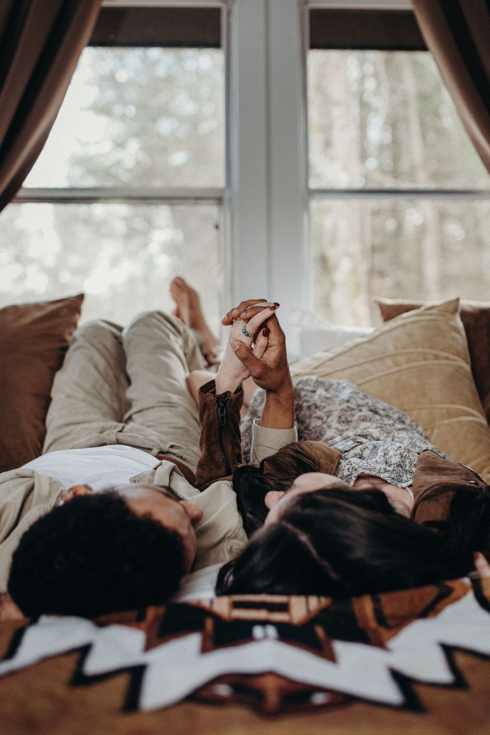 Two people lying down together on a couch by a bright window in a cozy setting.
