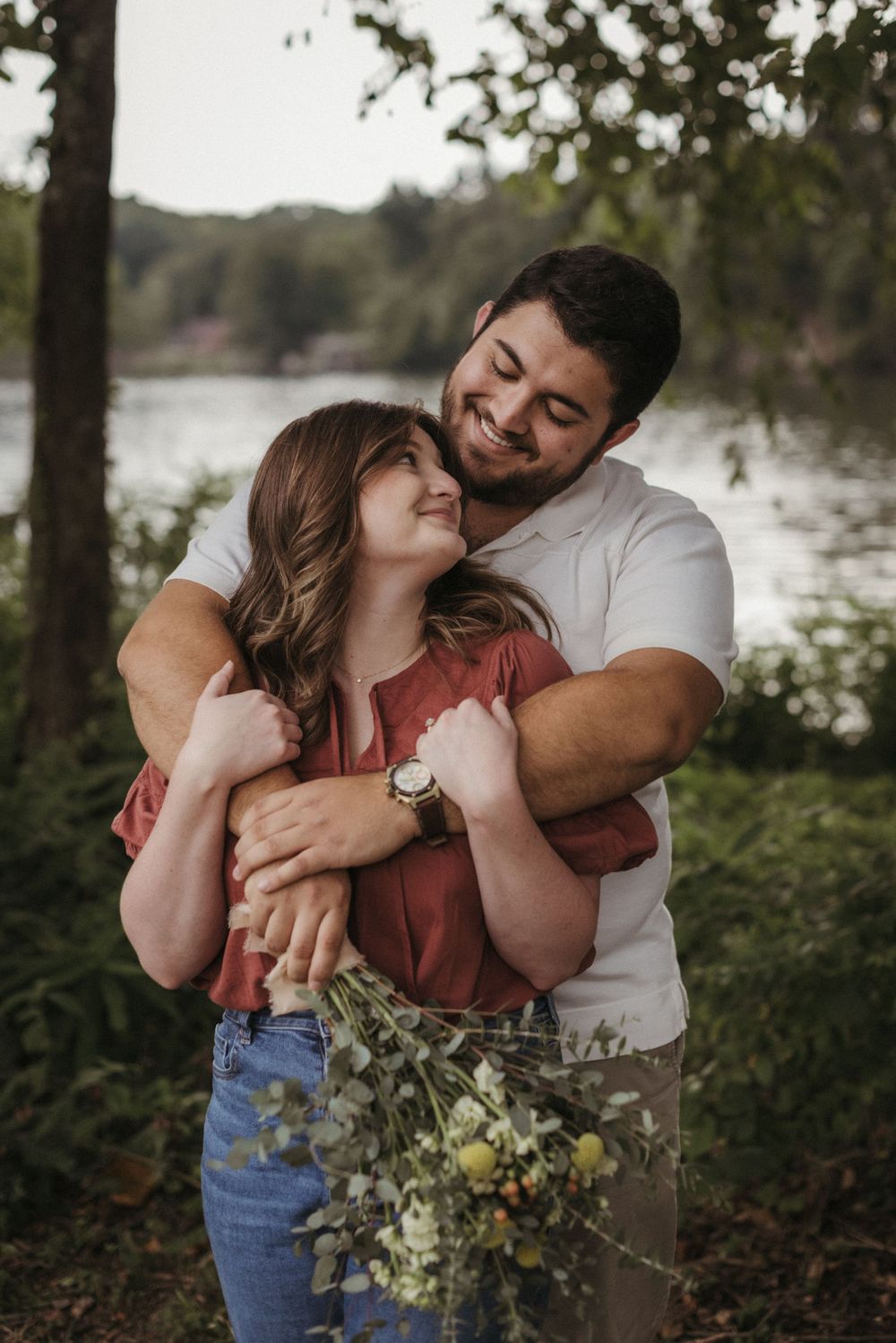 A romantic couple embrace while holding wildflowers by a lake at sunset.