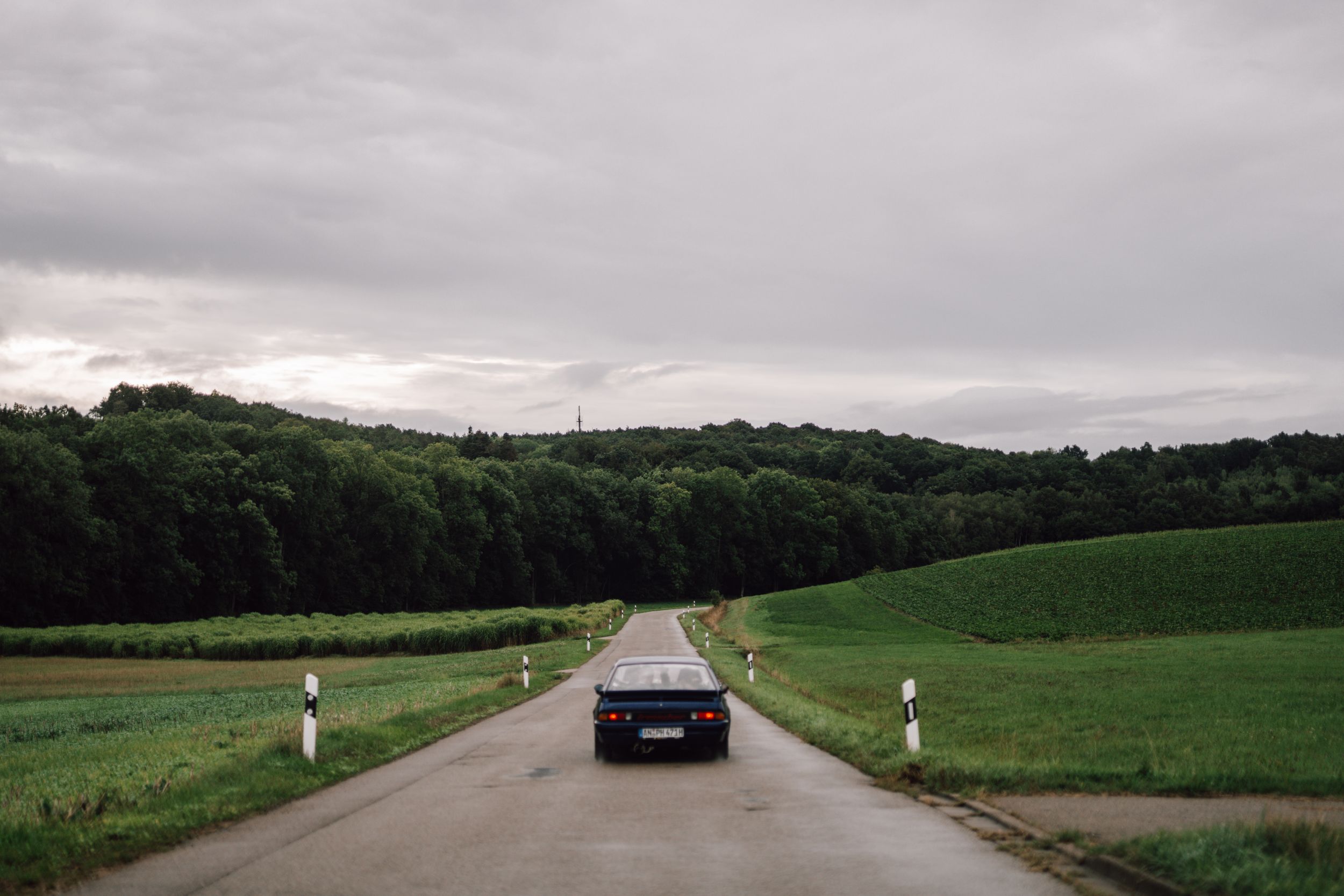 Car driving on rural road through green countryside under cloudy sky.