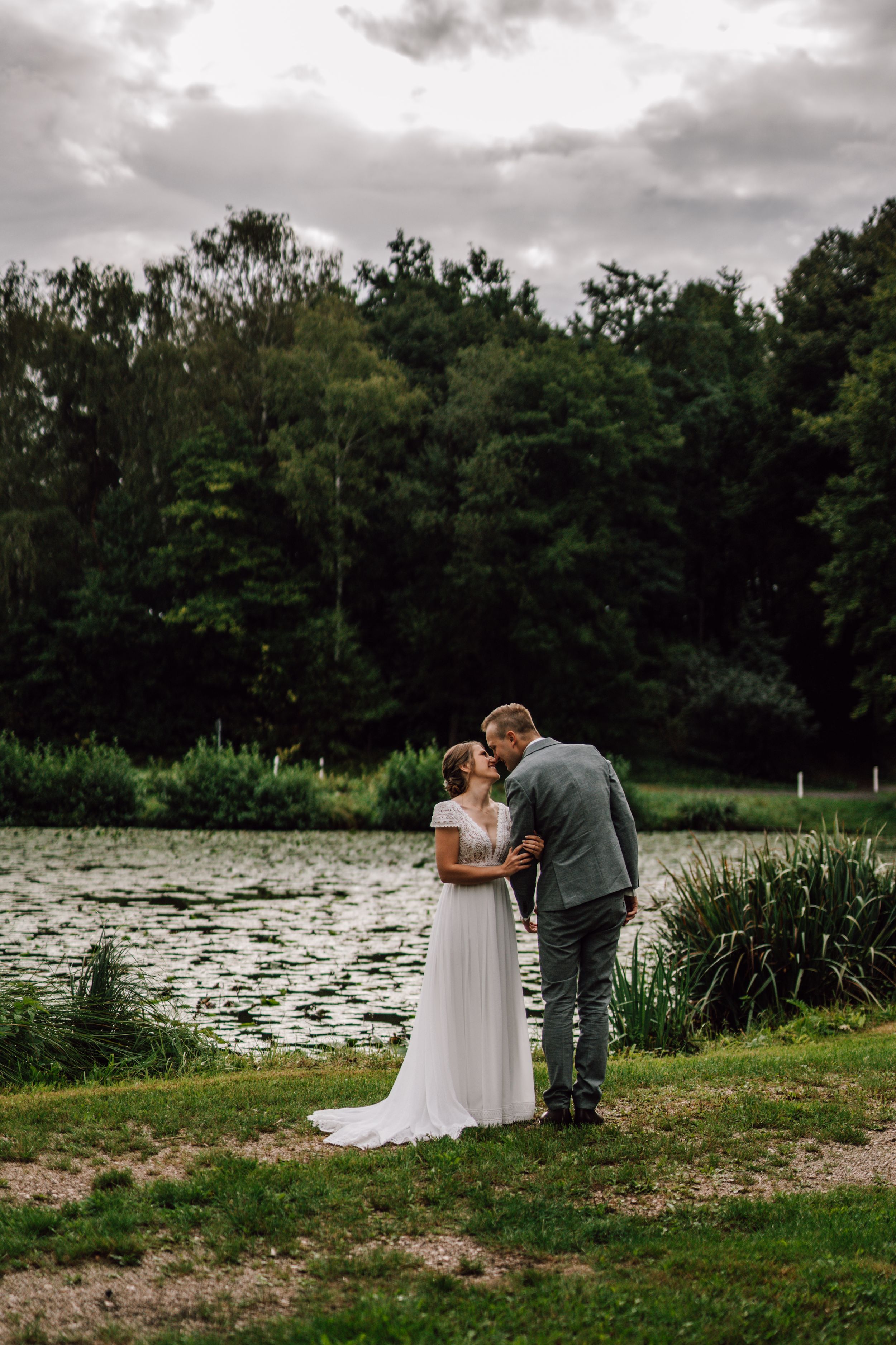 Newlyweds share an intimate moment by a scenic pond surrounded by lush greenery.