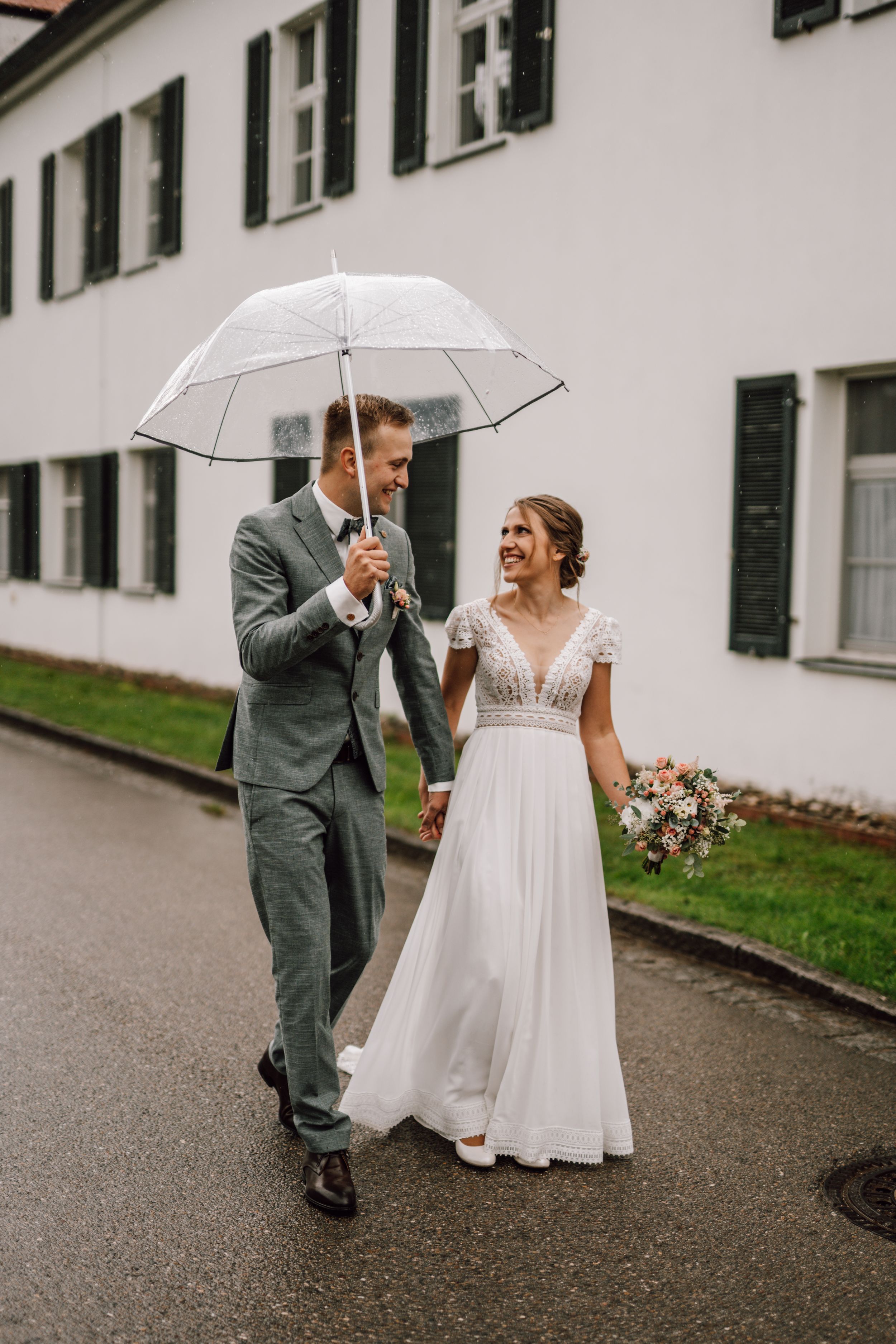 Couple walking in the rain under clear umbrellas against white building backdrop.