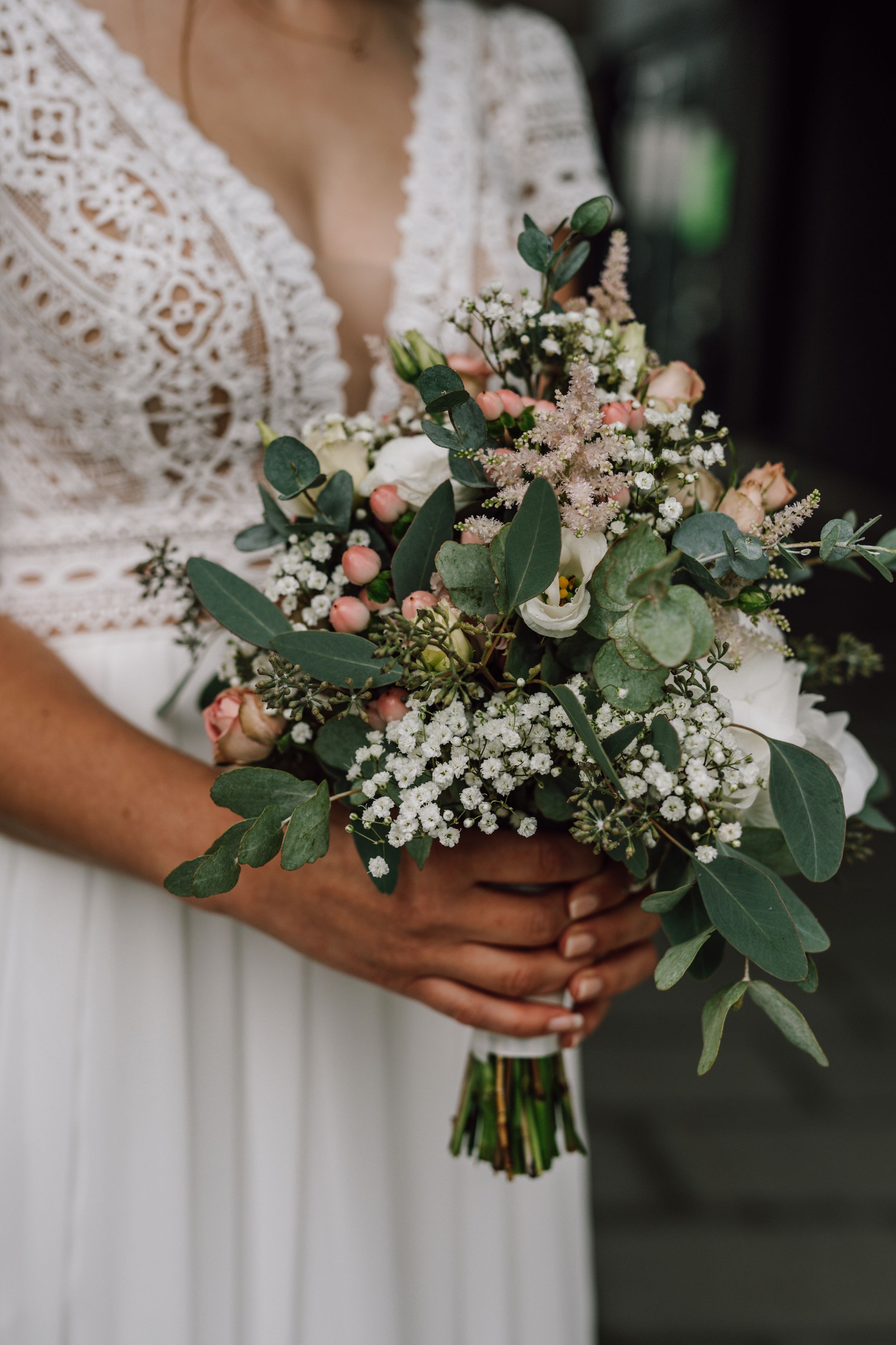Elegant bridal bouquet with eucalyptus, baby's breath, and blush roses held against a white lace wedding dress.
