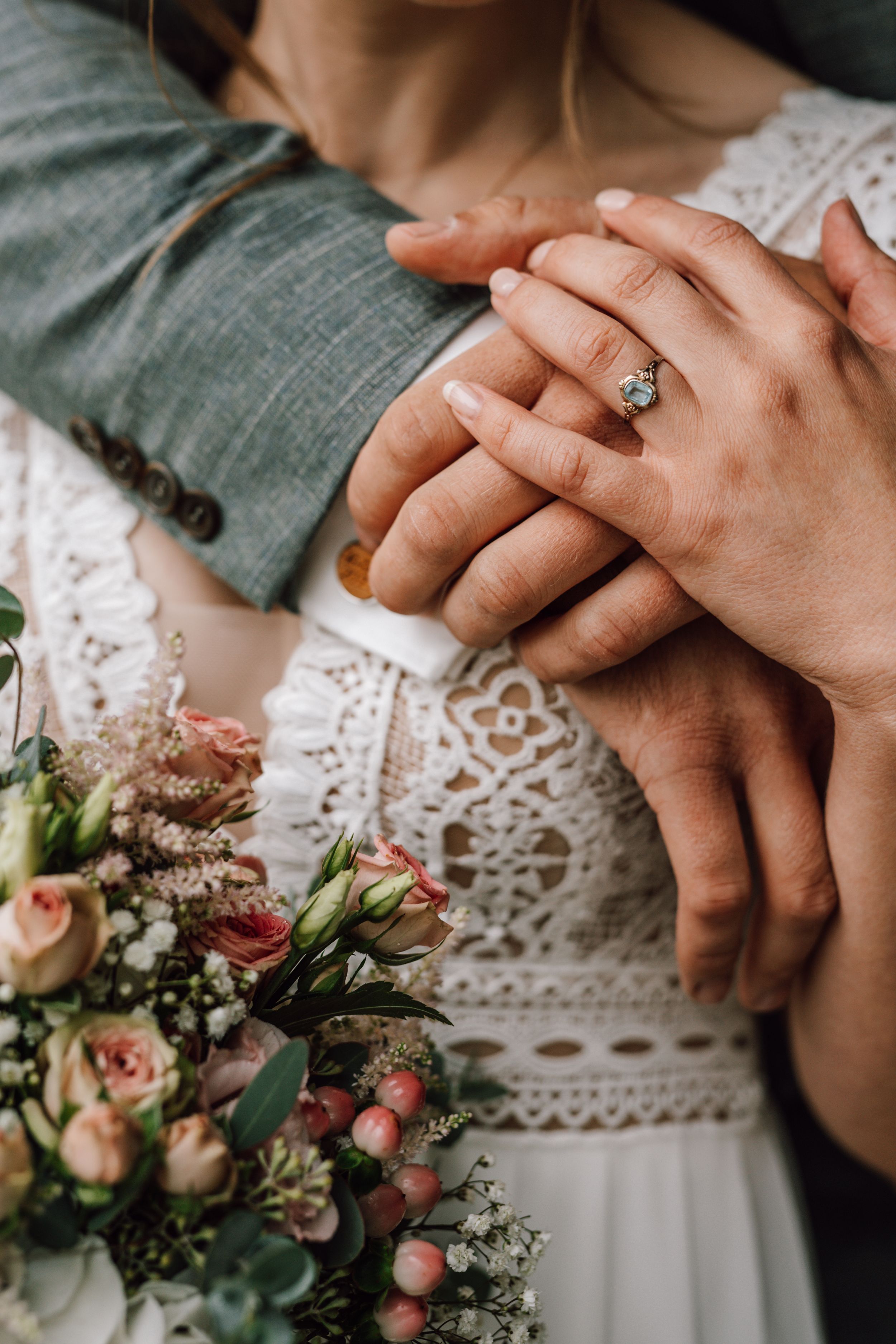 Close-up of hands with engagement ring and delicate lace wedding dress detail next to a soft pink floral bouquet.