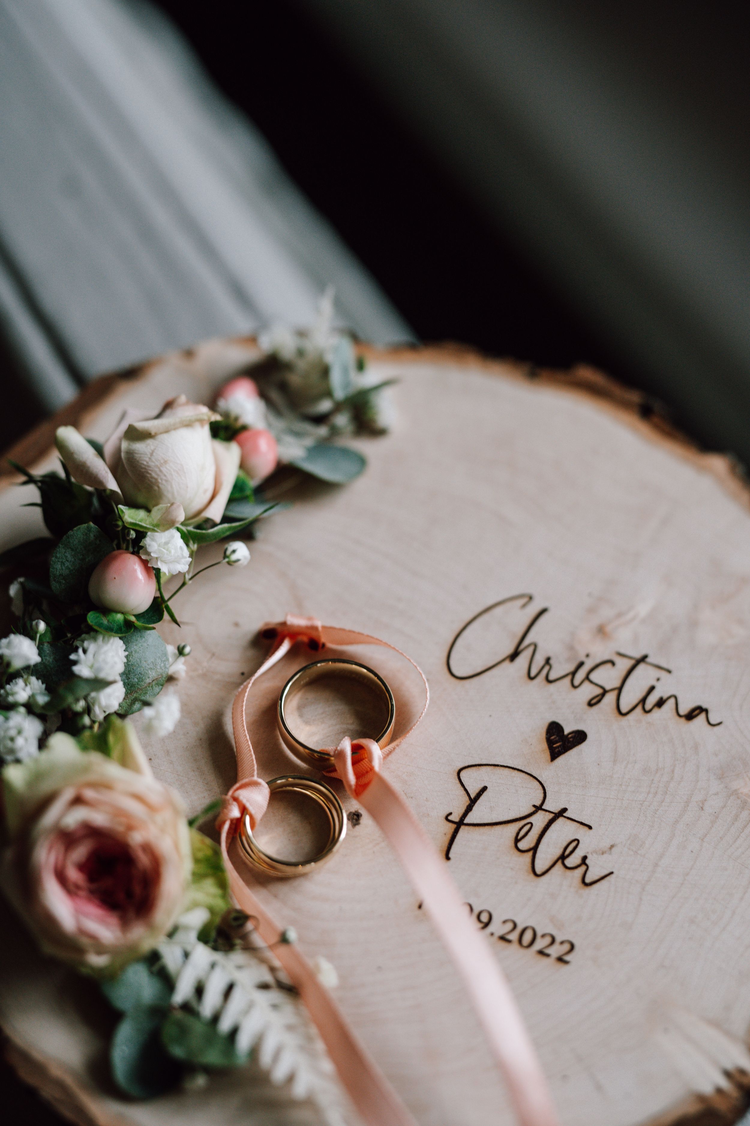 Wedding rings and floral decorations on wooden slice with engraved names and date.