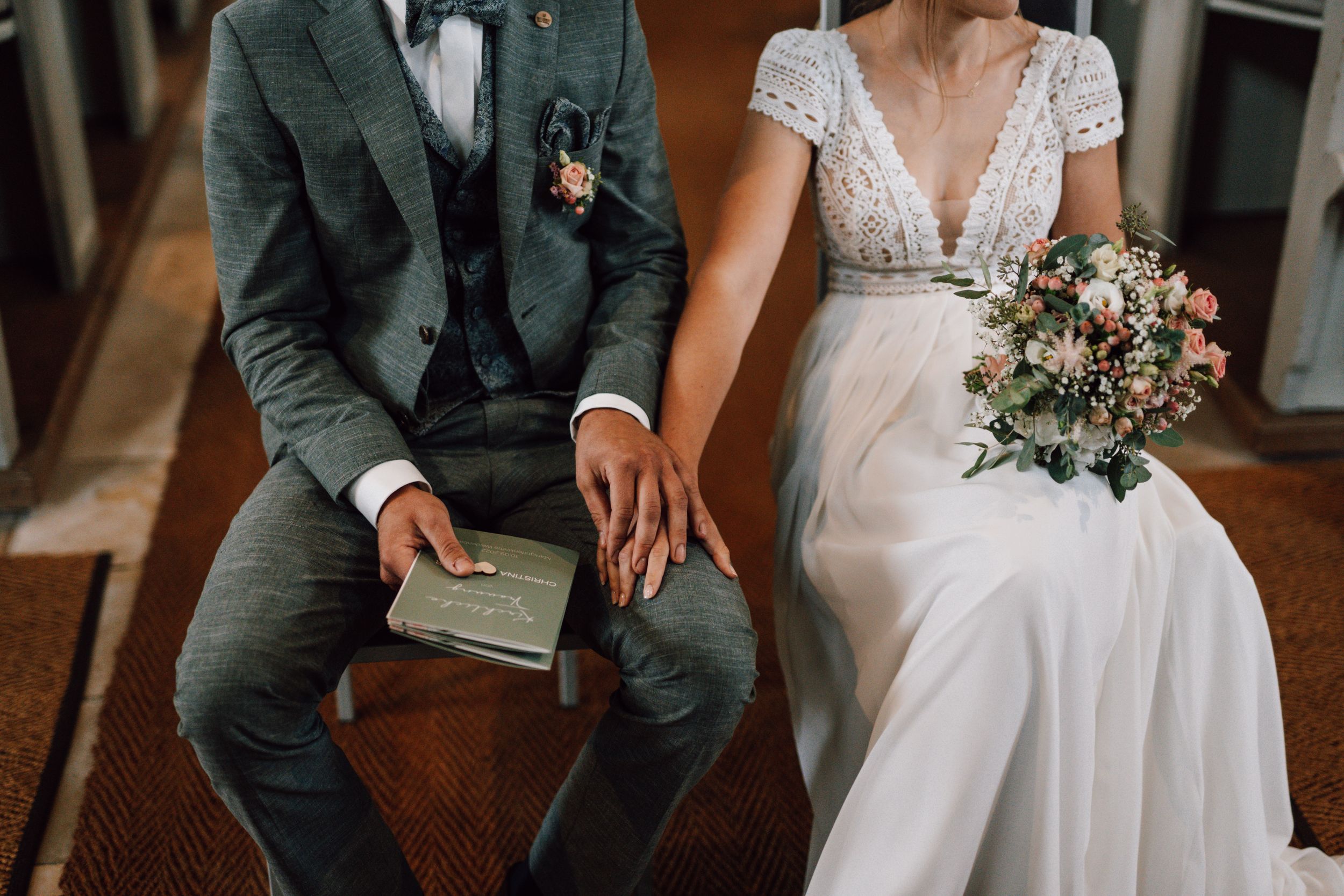 Couple sitting together in church, groom in grey suit and bride in lace white dress.