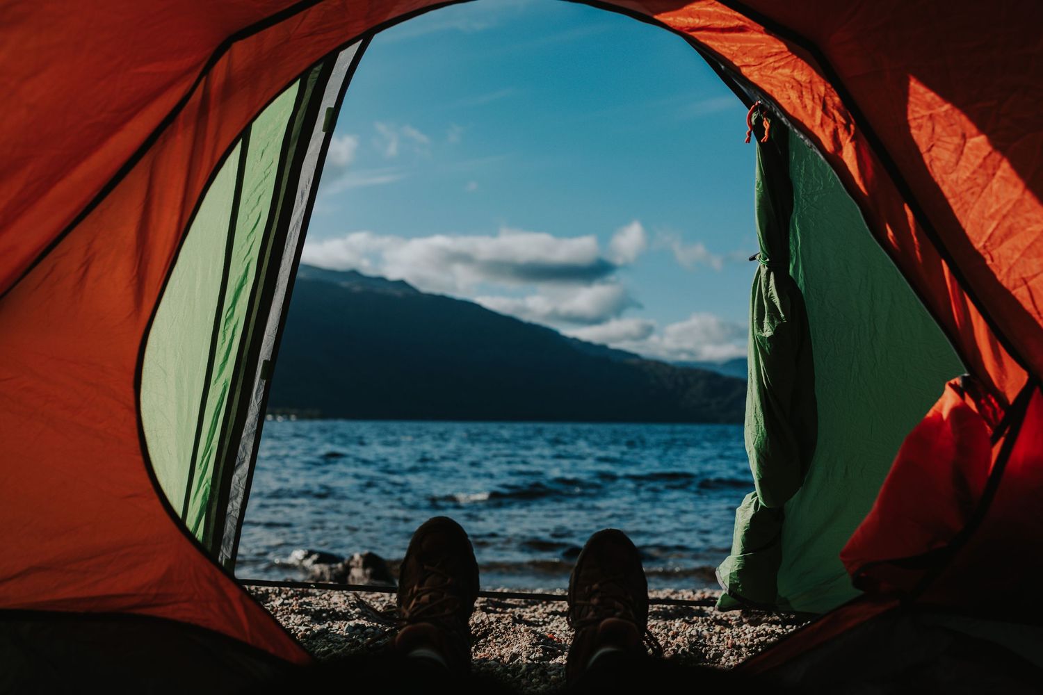 View through orange tent opening of lake and mountains with hiking boots visible.