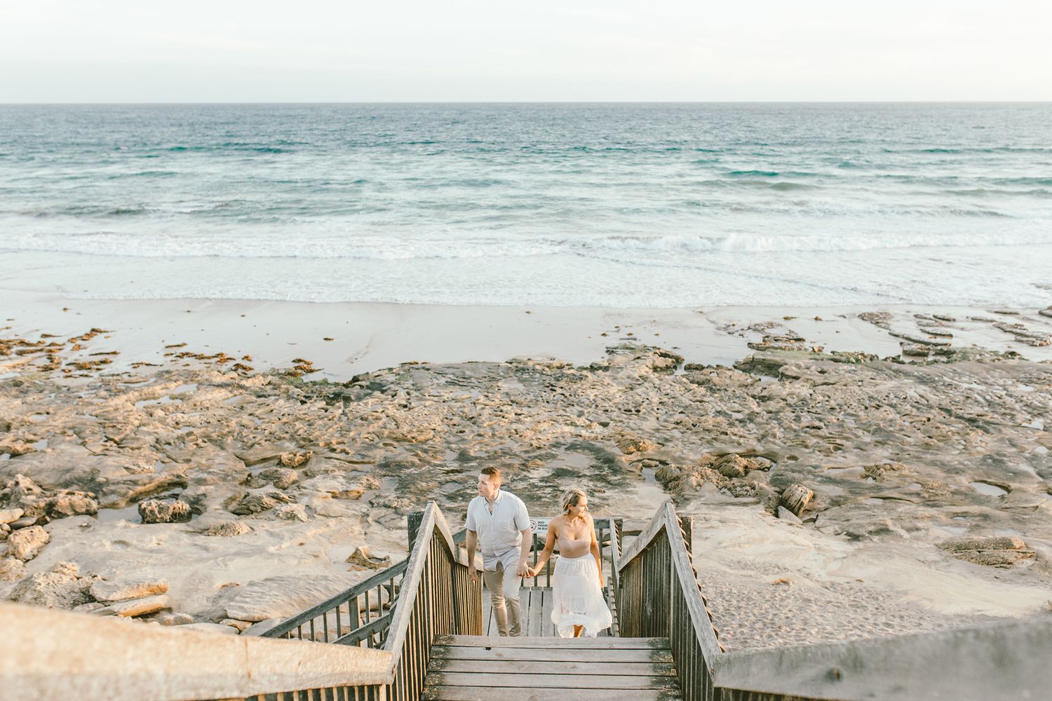 A couple descends wooden stairs leading to a rocky beach with ocean views.
