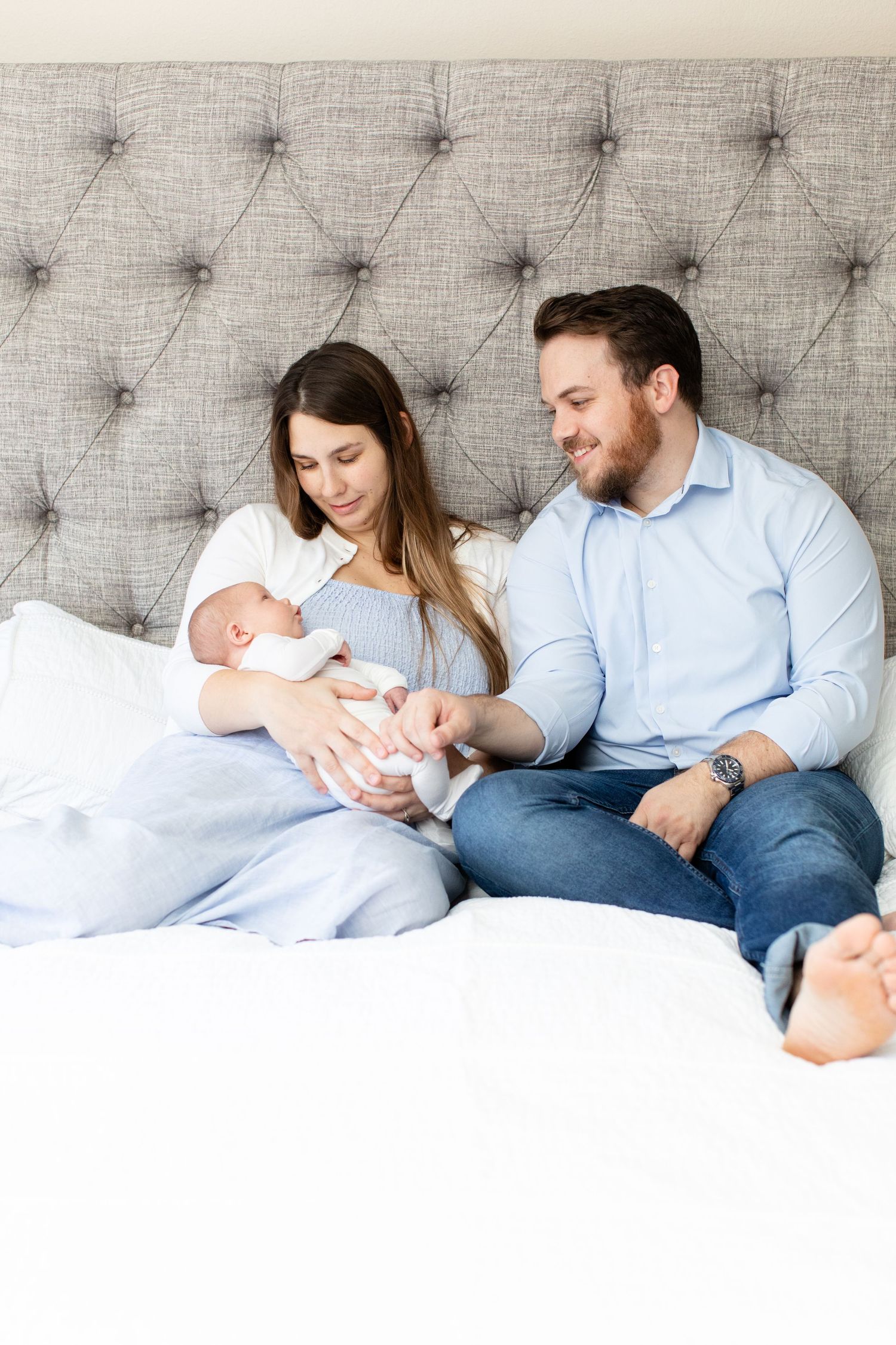 Parents cuddle with newborn baby on tufted grey headboard bed with white bedding.