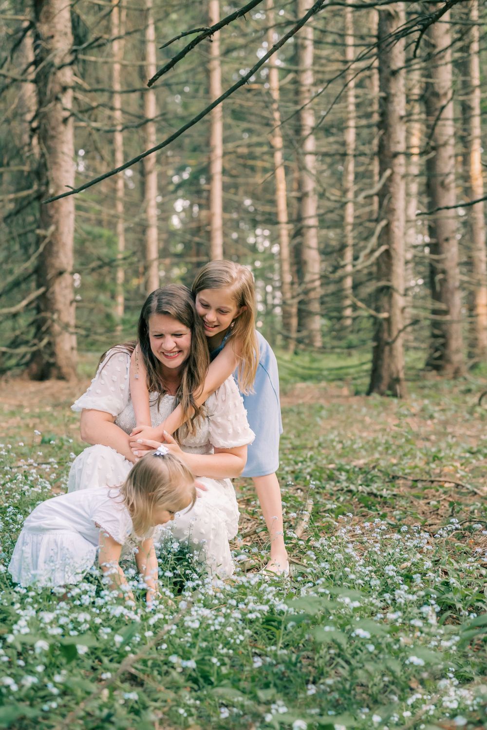 Three figures in white dresses explore a serene forest floor covered in small white flowers.