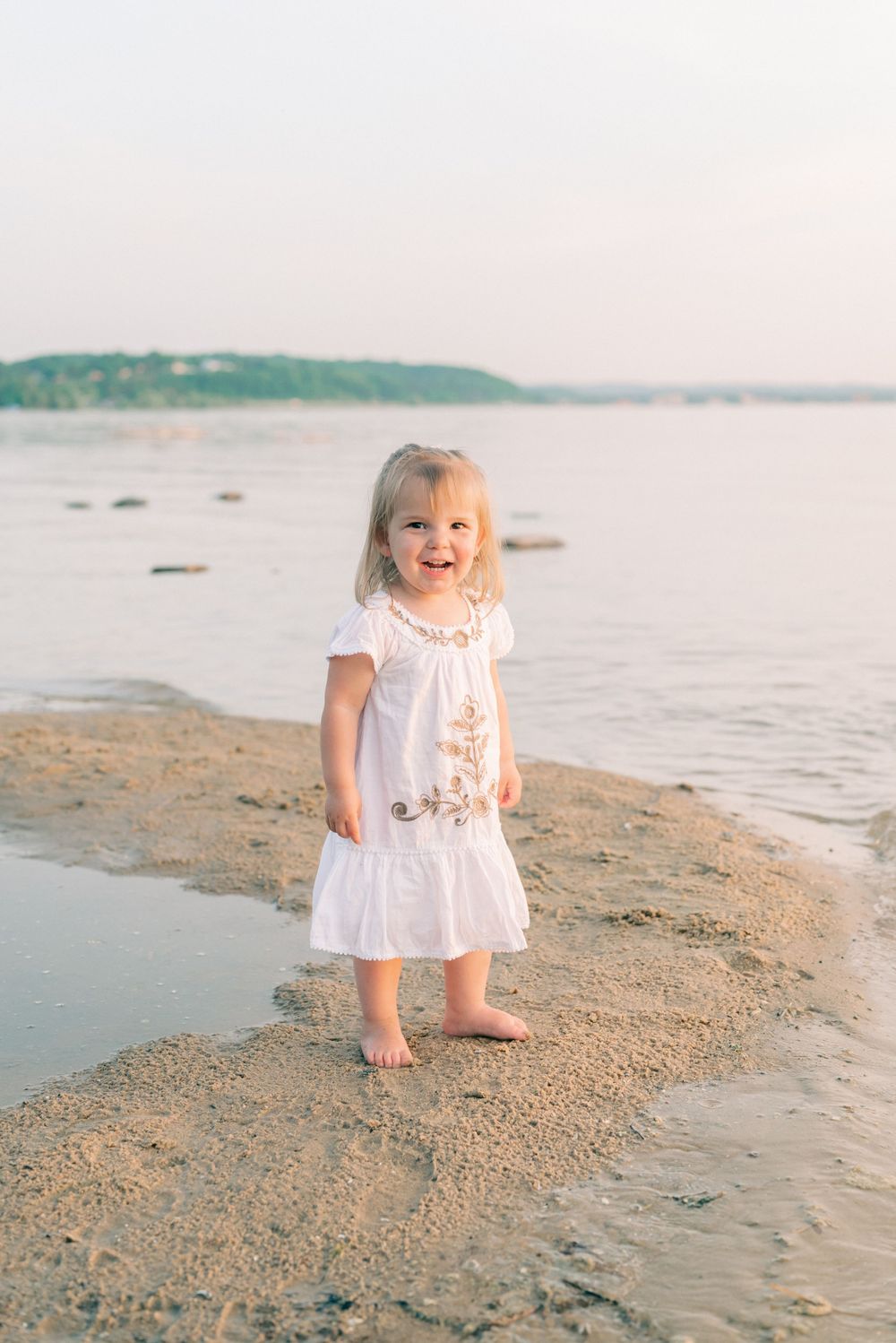 A small child in a white dress stands joyfully at the water's edge on a sandy beach.
