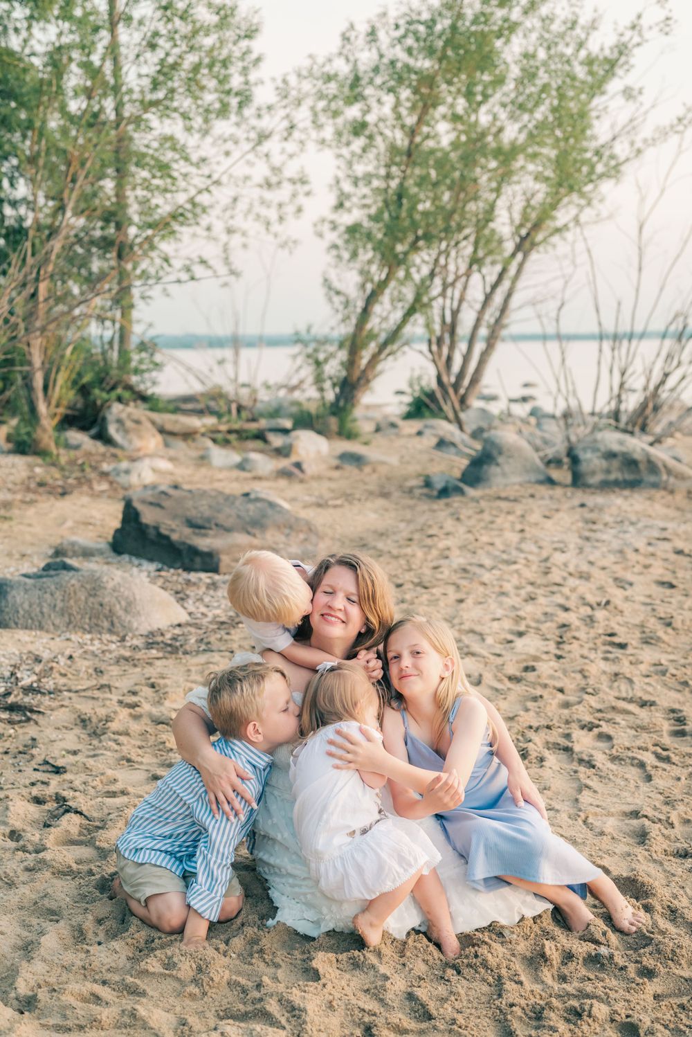 Group embracing on sandy beach with trees and water in background.