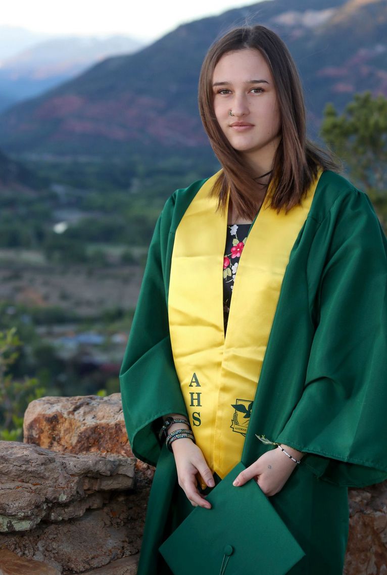 Graduate in green and gold academic regalia poses outdoors with mountains in background.
