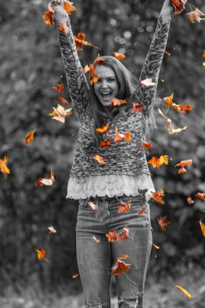 Person joyfully throwing fall leaves while wearing sweater and jeans with orange leaves in selective color.