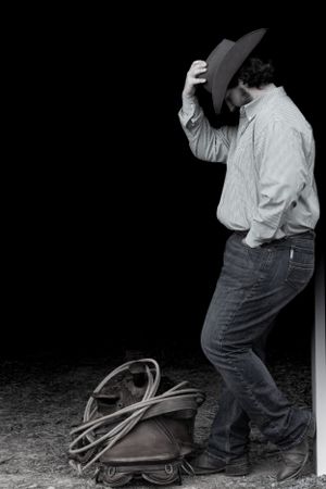 Figure in black western attire tipping cowboy hat against dark background with rope on ground.