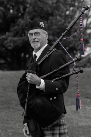 Bagpiper in traditional Scottish dress smiling while holding decorated bagpipes outdoors.