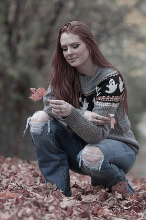Person in patterned sweater and ripped jeans examining autumn leaves while crouching outdoors.