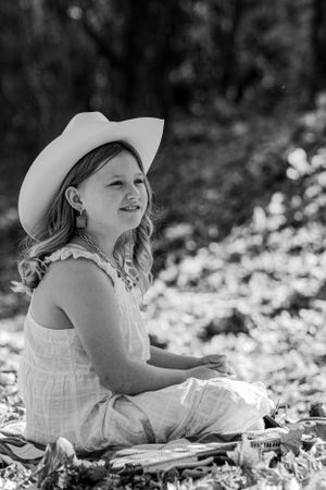 Young person in white dress and cowboy hat sitting on ground in black and white portrait.
