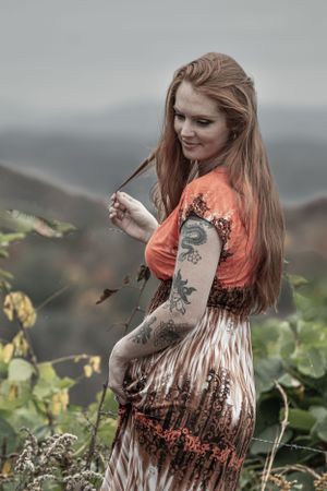 Person in orange top and patterned dress showing tattoos while posing on hillside.