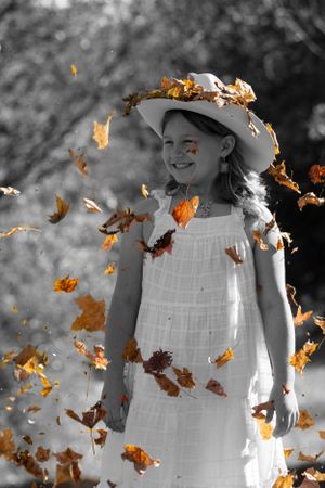 Selective color photo showing orange leaves swirling around someone in white dress and sun hat.