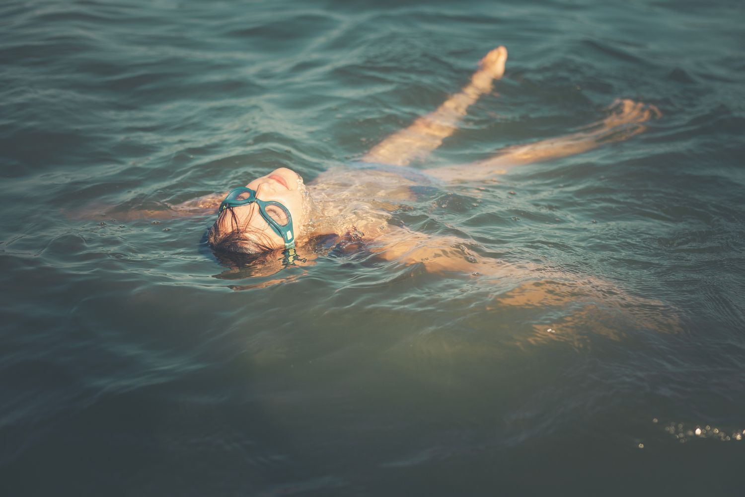 A child floats peacefully on their back in calm ocean water, wearing blue goggles and basking in the warm sunlight.