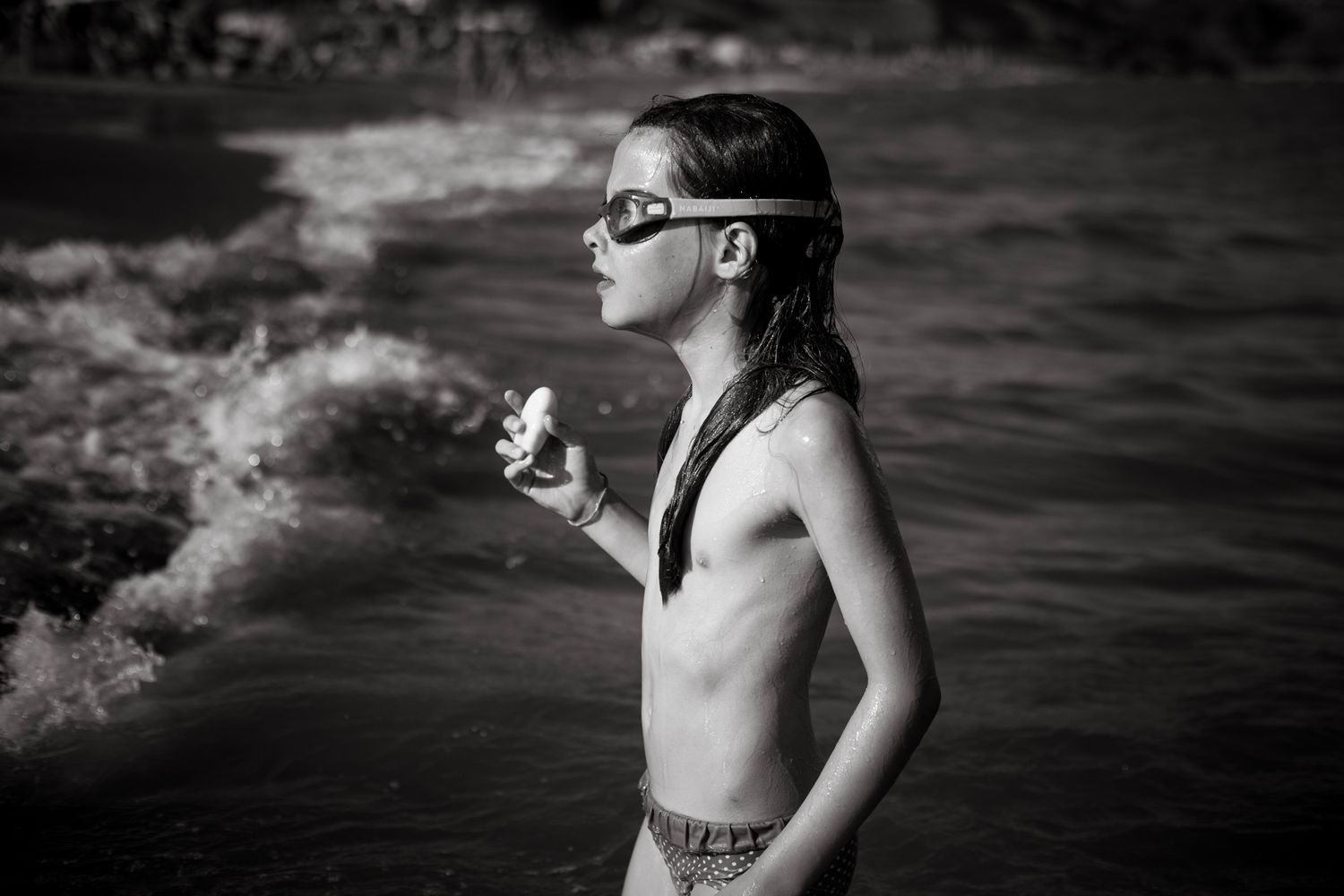 A young girl wearing swim goggles stands at the ocean's edge, gazing out at the waves in a thoughtful black-and-white beach scene.
