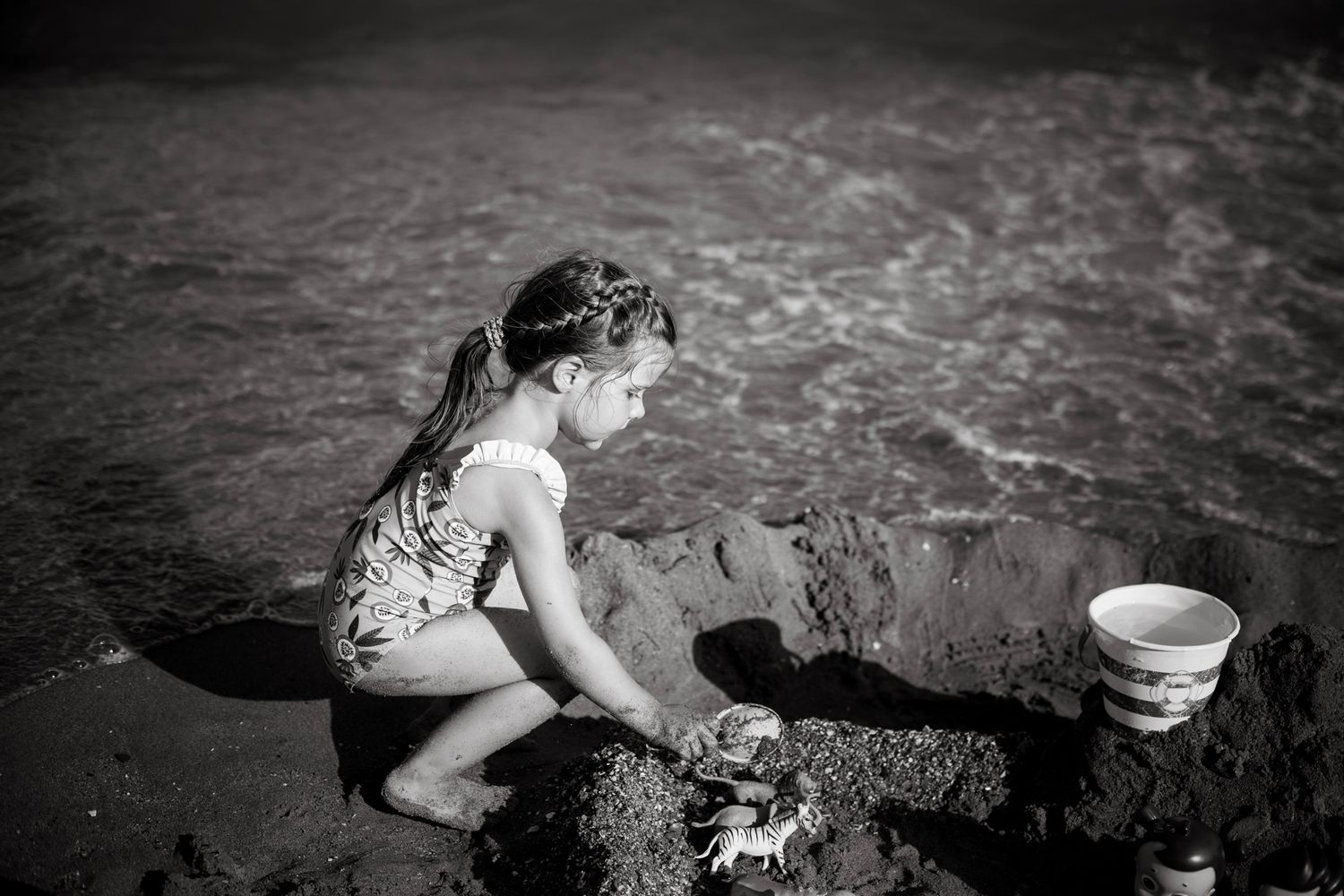 A young girl kneels in the sand by the shoreline, deeply focused as she builds with toys beside a bucket in this serene black-a.