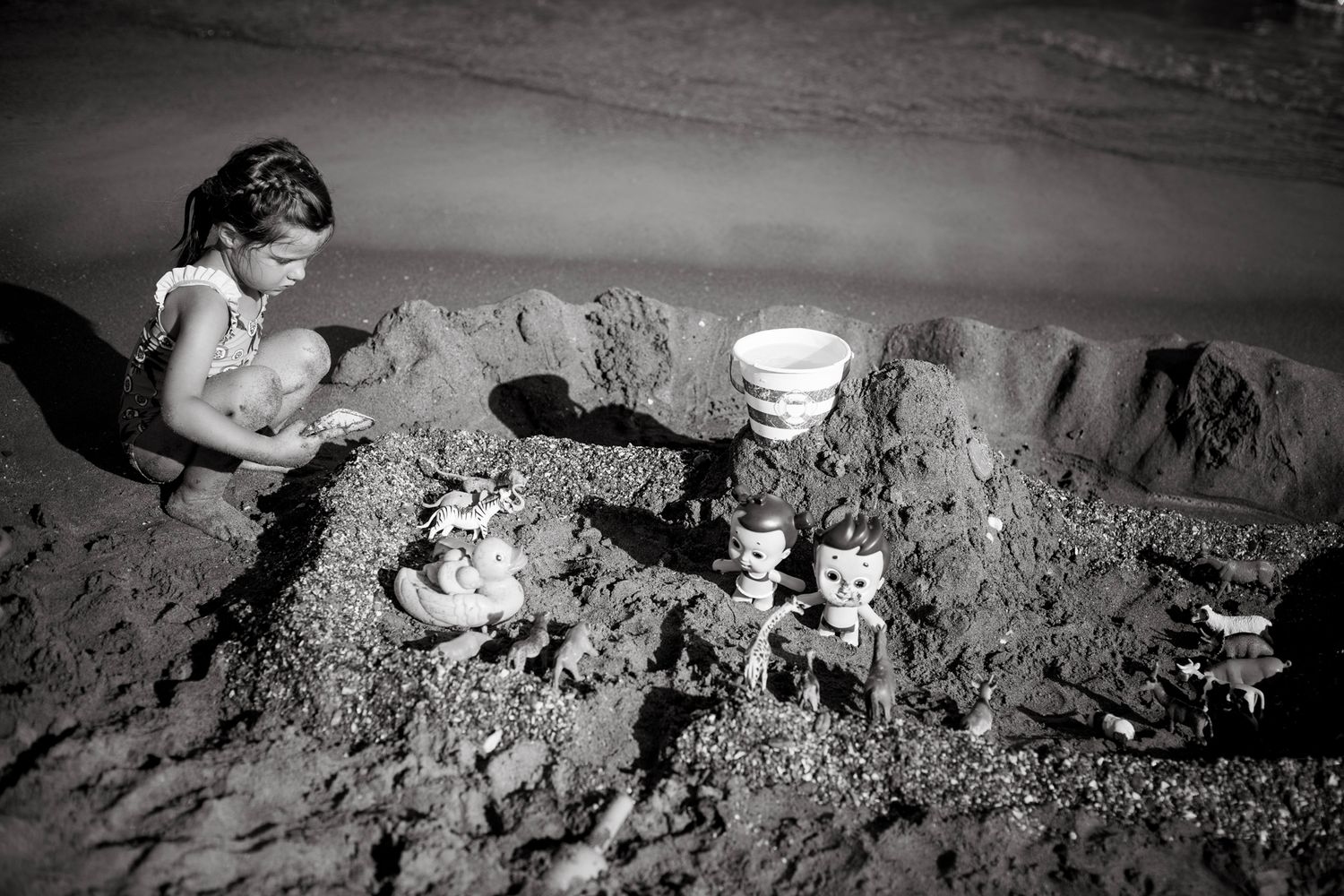 A young girl crouches beside an elaborate sandcastle decorated with toy figures and a bucket, fully absorbed in her beachside castle.