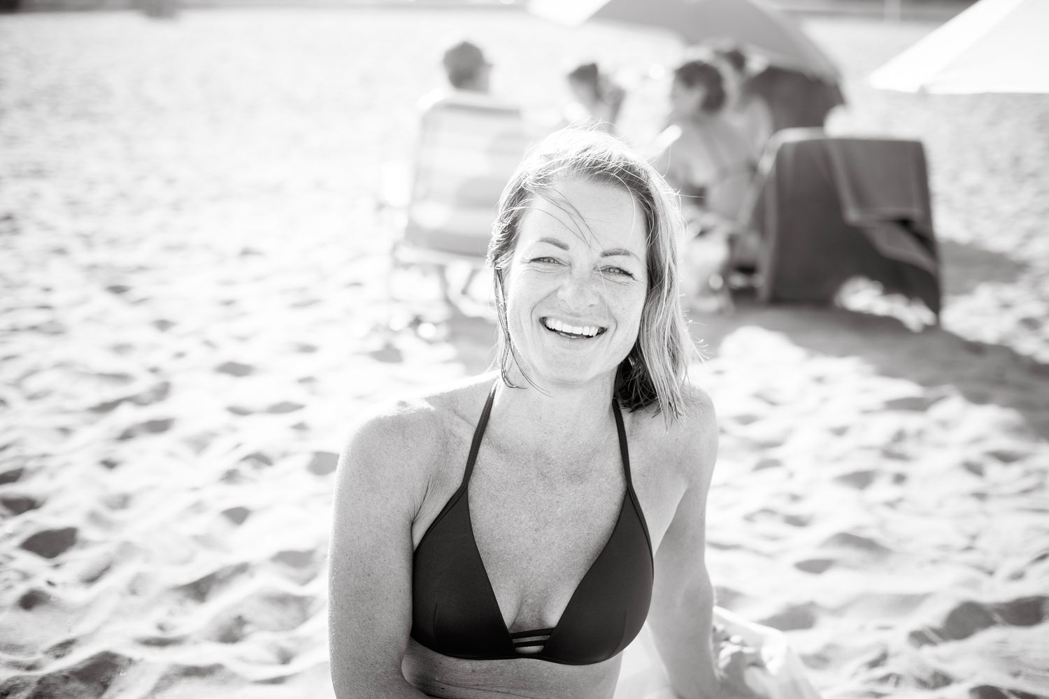 A smiling woman in a bikini top sits on the sandy beach, enjoying a sunny day in the background  of umbrellas.