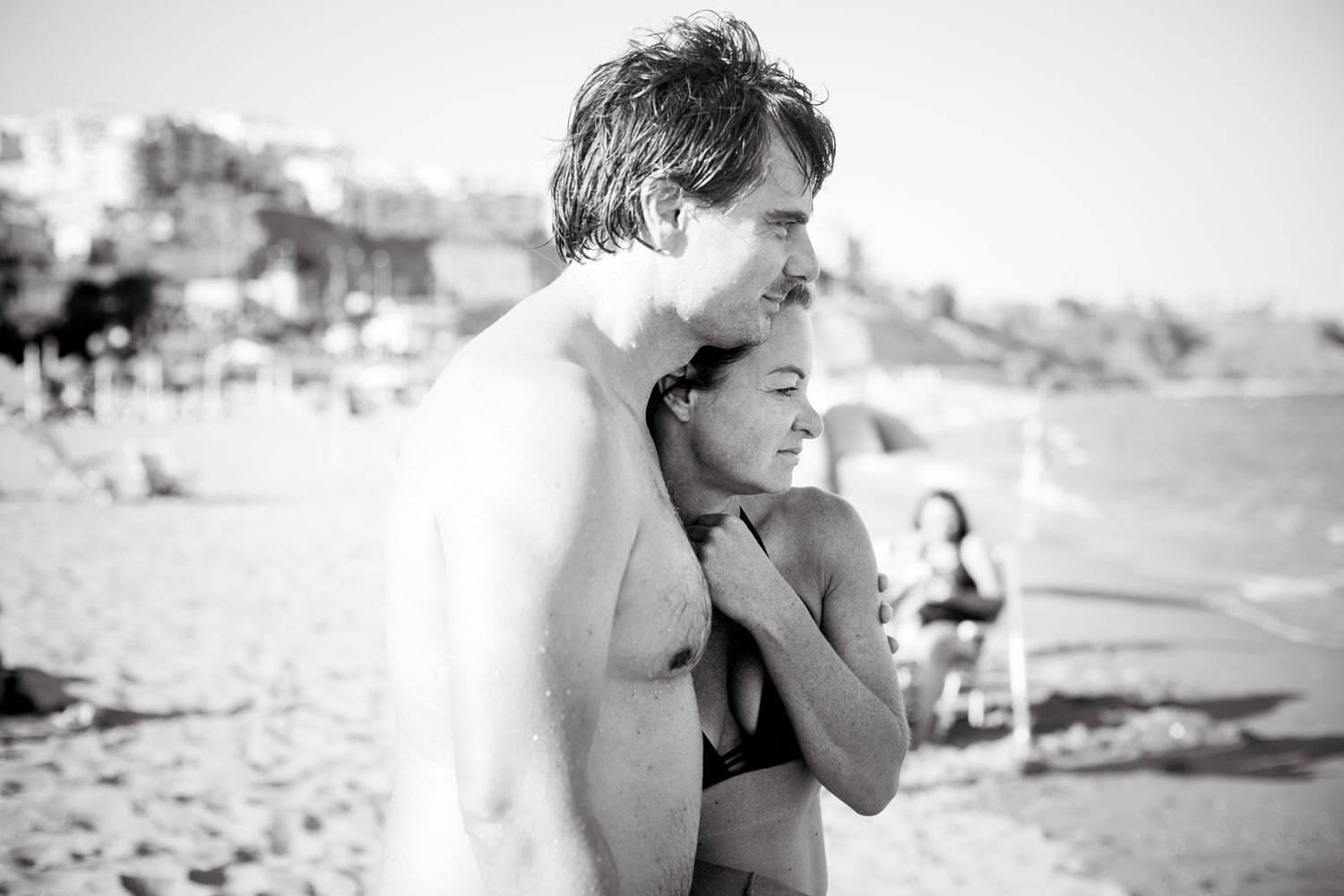 A couple stands close together on the beach, gazing out at the sea and sharing a quiet, affectionate moment in black and white.