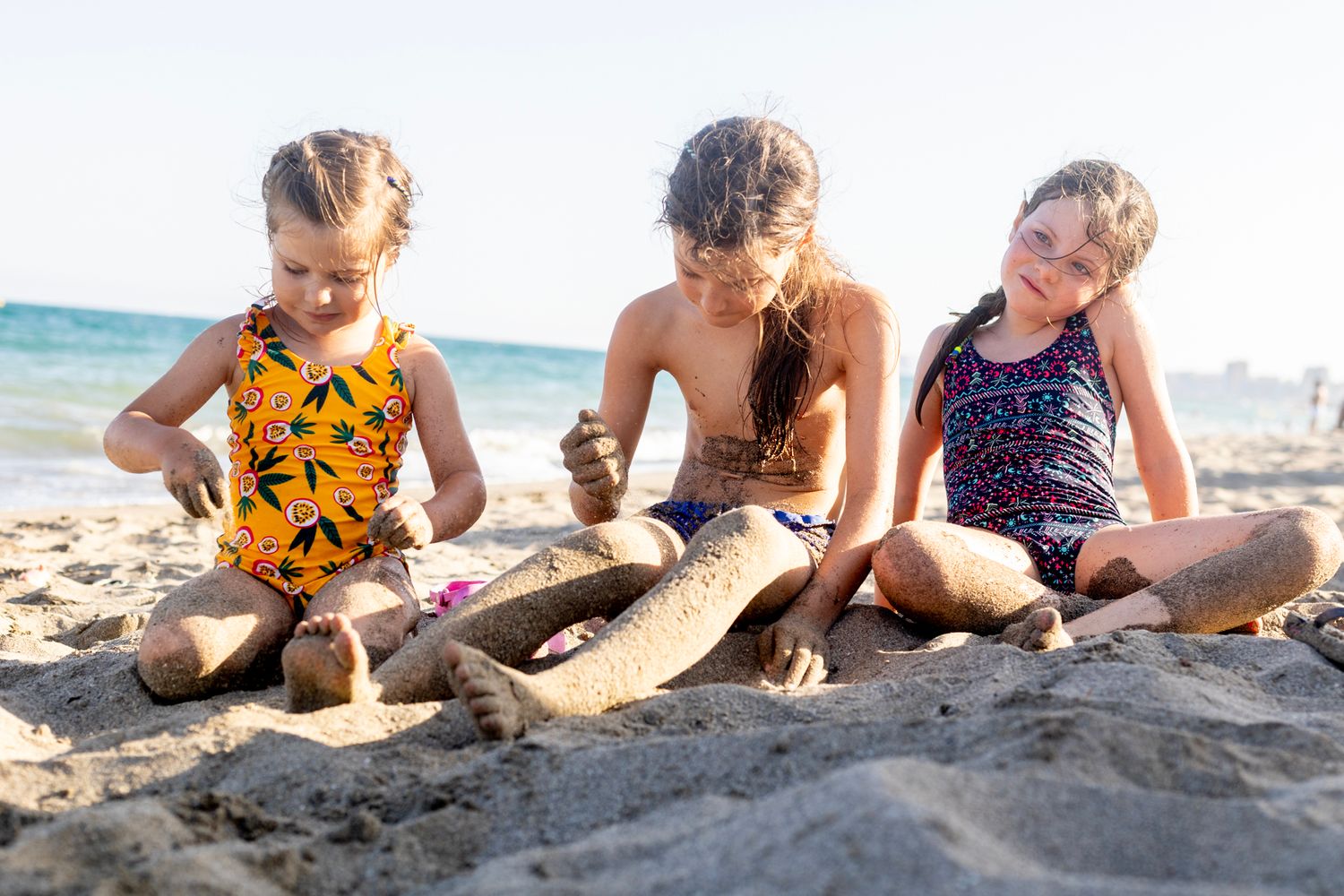 Three girls sit on the sandy beach, focused and playful as they cover their legs in sand under the bright afternoon sun.
