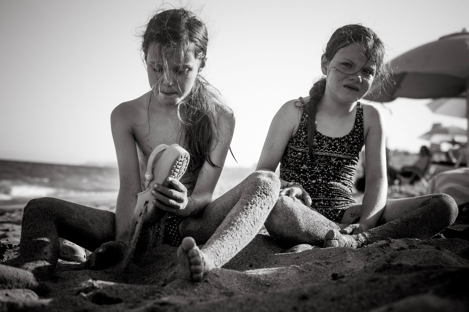 Two girls sit in the sand at the beach, one focused on brushing a doll's hair while the other looks on, both covered in sand in a candid.