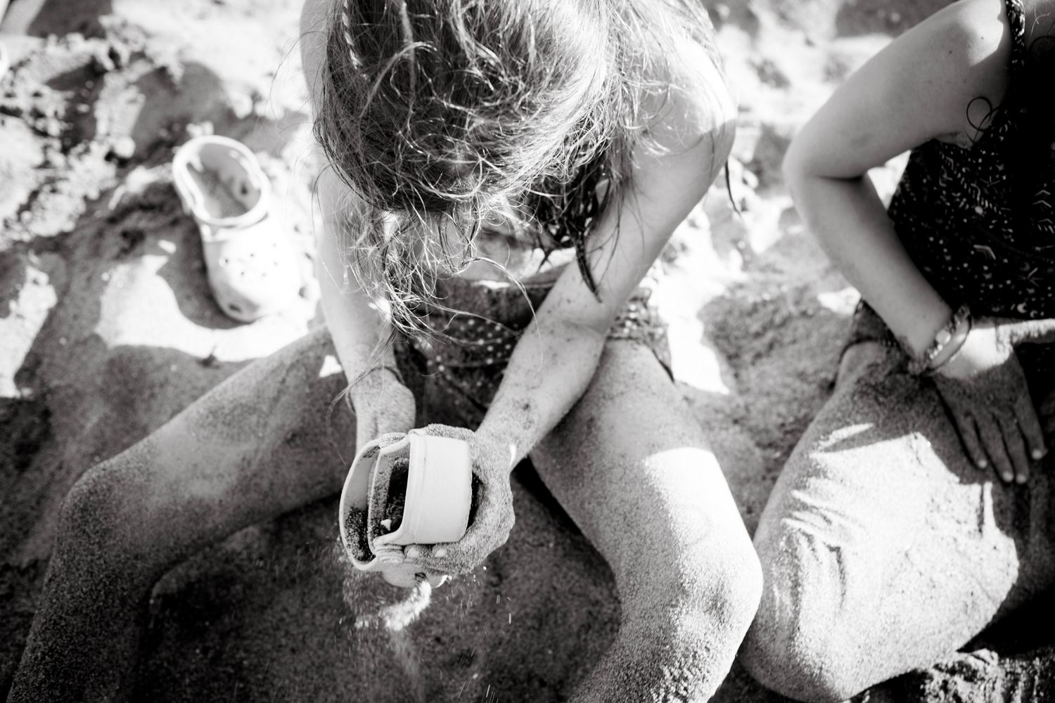 A sandy child examines a small container on their lap while sitting crosslegged on the beach, fully engaged in their discovery under the sun