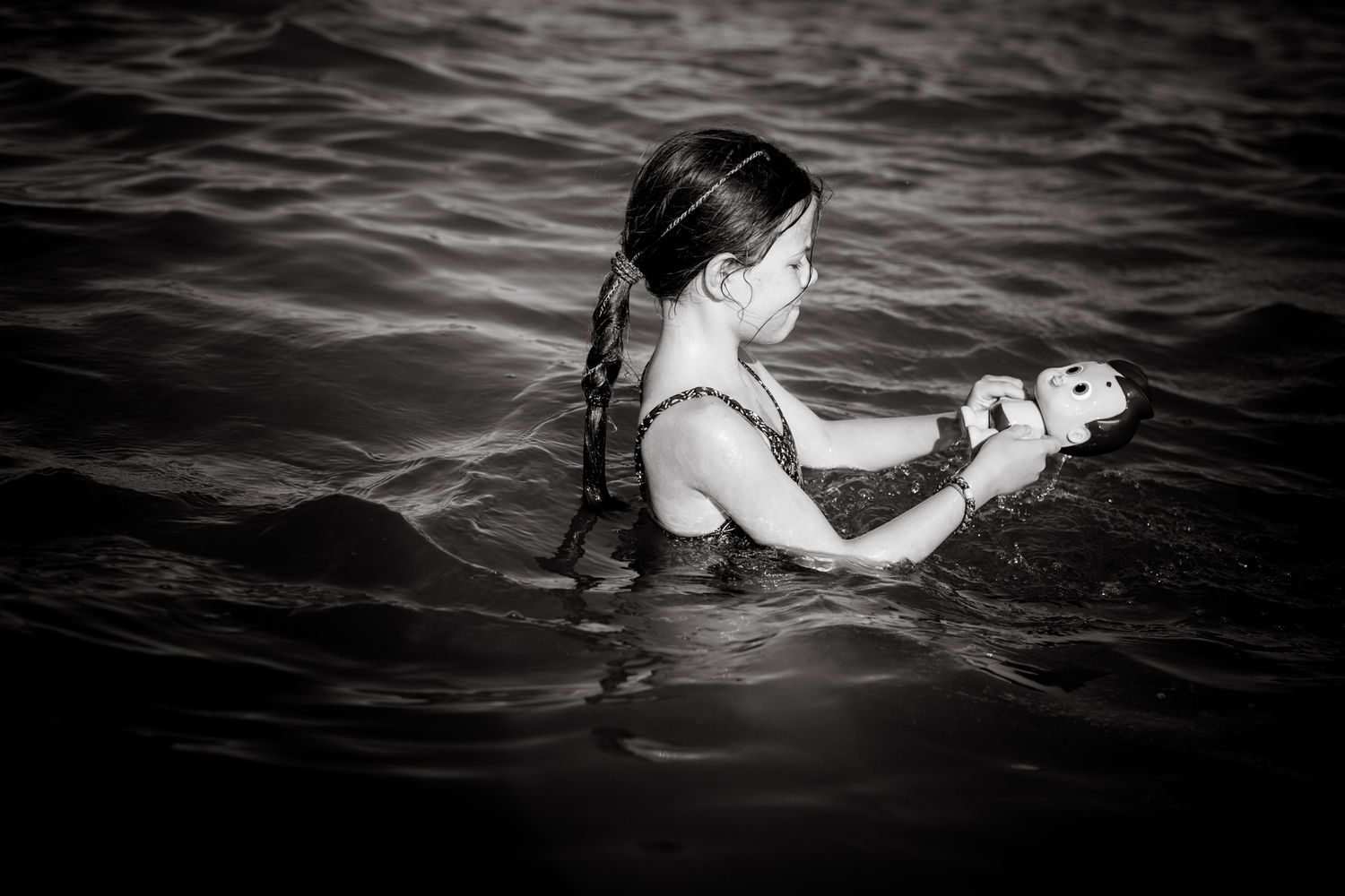 A girl with braided hair smiles while holding a toy doll in the water, captured mid-play in a calm, black-and-white ocean scene.