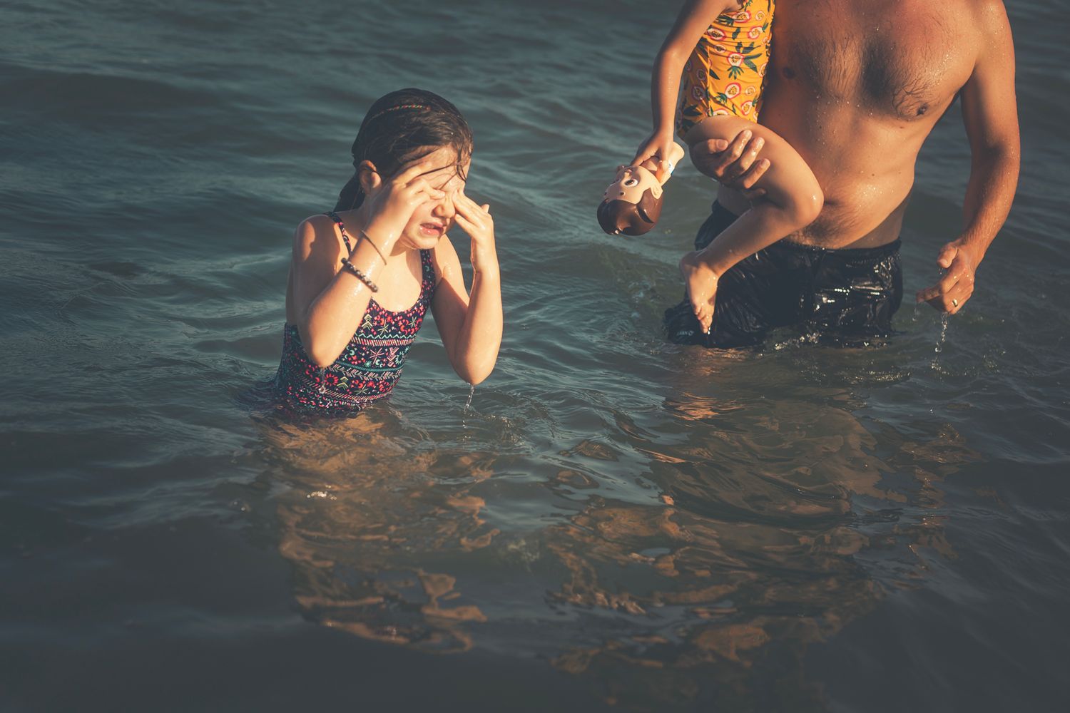 A girl wipes water from her face while standing in the ocean, beside her father holding her brother during a sunny beach outing.