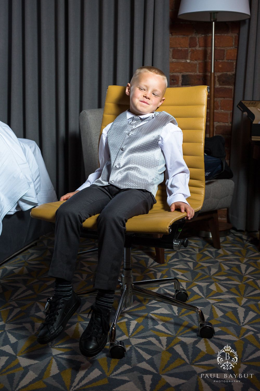 Young formal attire on yellow chair against grey curtains and brick wall.