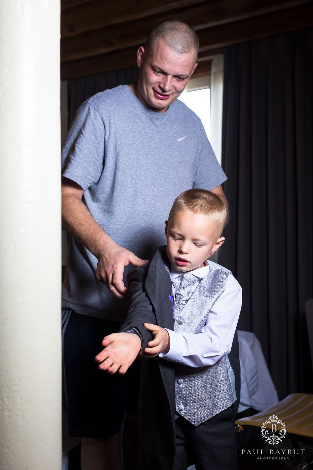 A man in a grey t-shirt helps a young boy in formal attire adjust his outfit in an indoor setting.